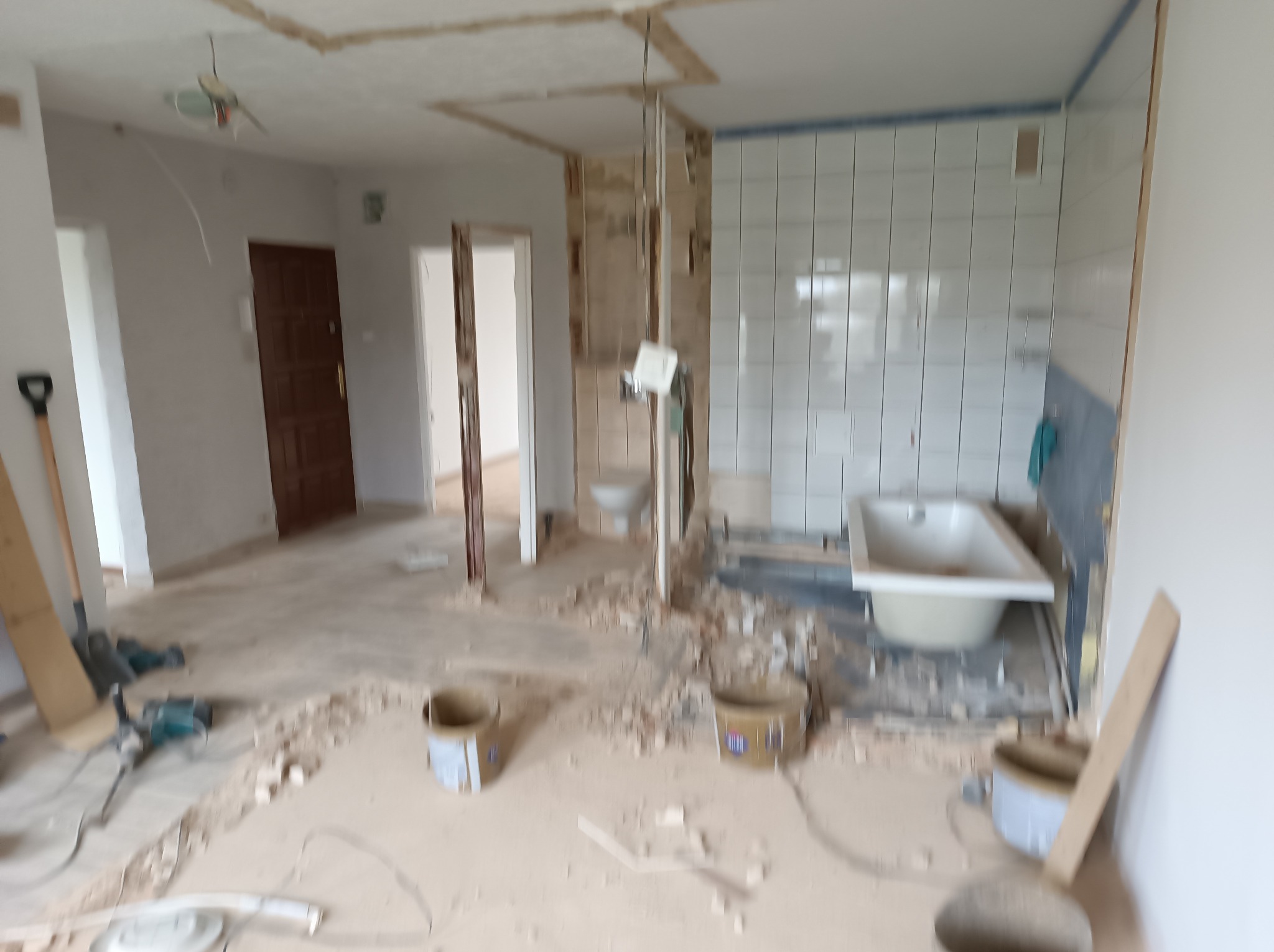 Wnętrze w trakcie demontażu: usunięte płytki w łazience, gruz na podłodze, widoczna wanna, narzędzia i puste wiadra po farbie, odsłonięta instalacja elektryczna w suficie.