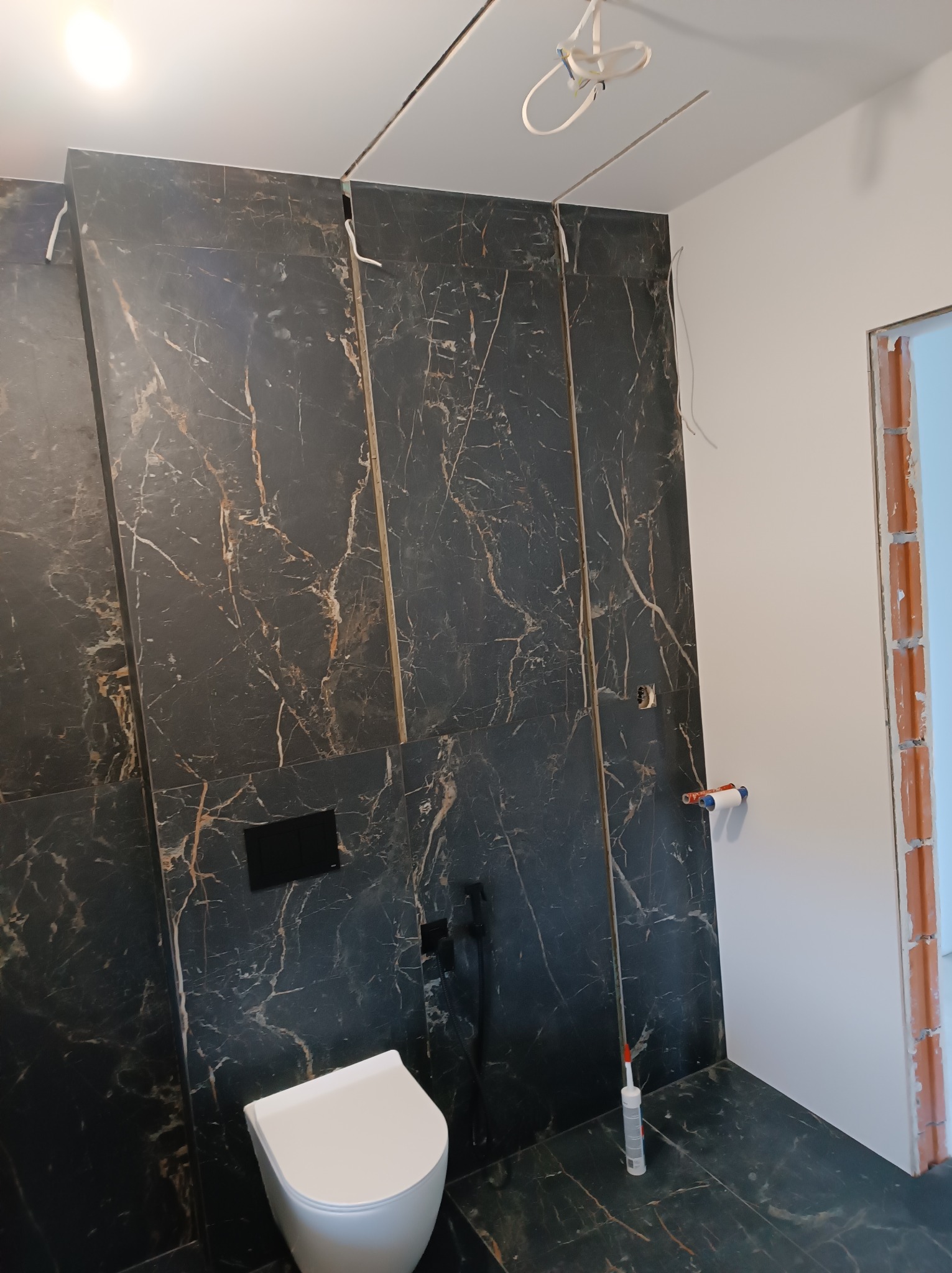 Niedokończona łazienka z czarnymi płytkami imitującymi marmur, wisząca toaleta i widoczne instalacje pod sufitem, silikon w tubie na podłodze i surowy otwór drzwiowy.