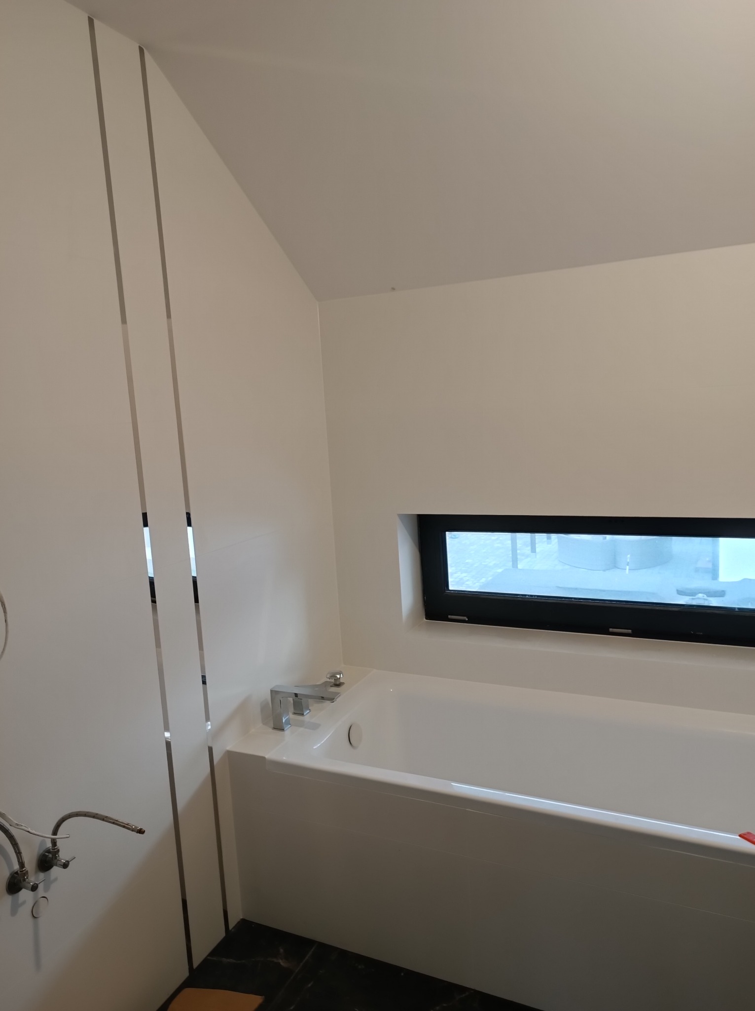 Nowoczesna biała łazienka z wanną, minimalistycznym oknem i czarną marmurową podłogą, widoczne rury instalacji wodnej przed zabudową.