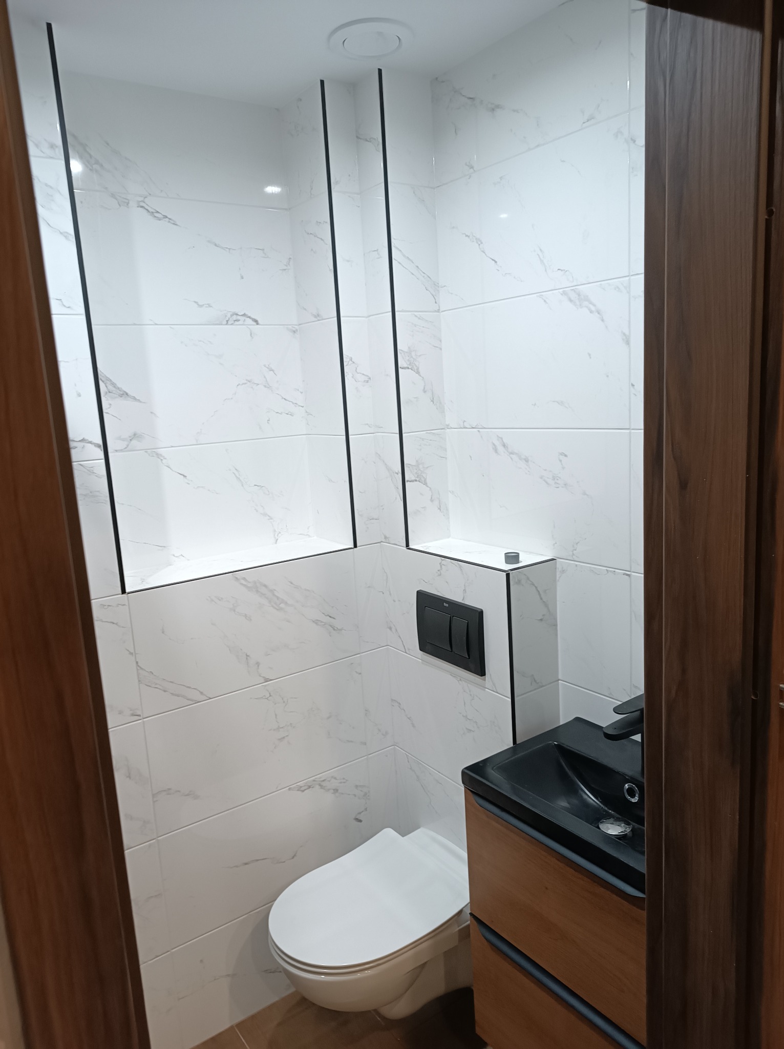 Nowoczesna toaleta w odcieniach bieli i czerni, z marmurowymi płytkami na ścianach, wbudowanymi niszami z czarnymi obramowaniami oraz umywalką z szafką w kolorze drewna.