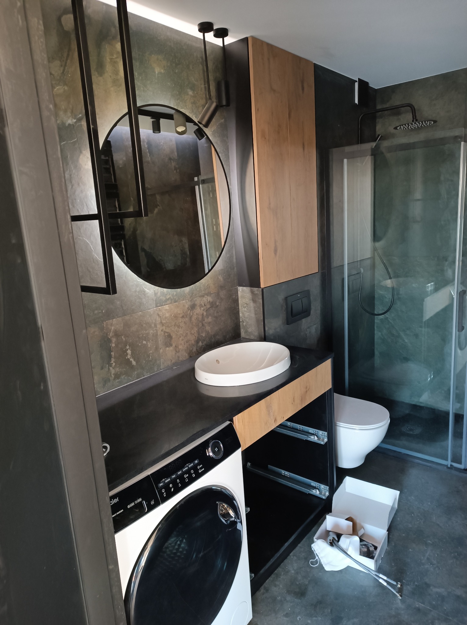 Nowoczesna łazienka w trakcie urządzania: pralka Haier, okrągłe lustro z oświetleniem, kabina prysznicowa, toaleta oraz akcesoria montażowe w otwartym pudełku na podłodze.