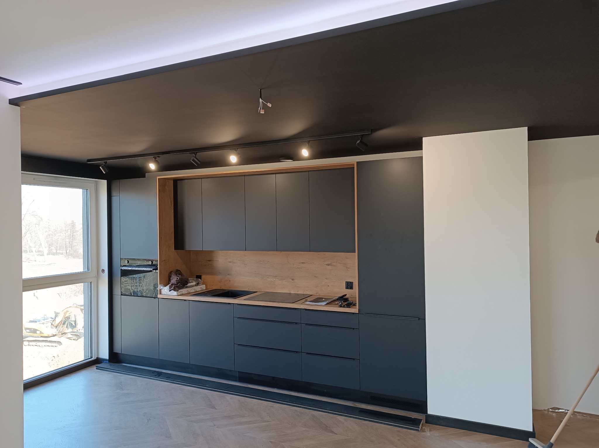 Nowoczesna kuchnia z ciemnymi, matowymi frontami szafek, drewnianym akcentem na ścianie i czarnym sufitem z oświetleniem szynowym; widok na plac budowy przez okno.