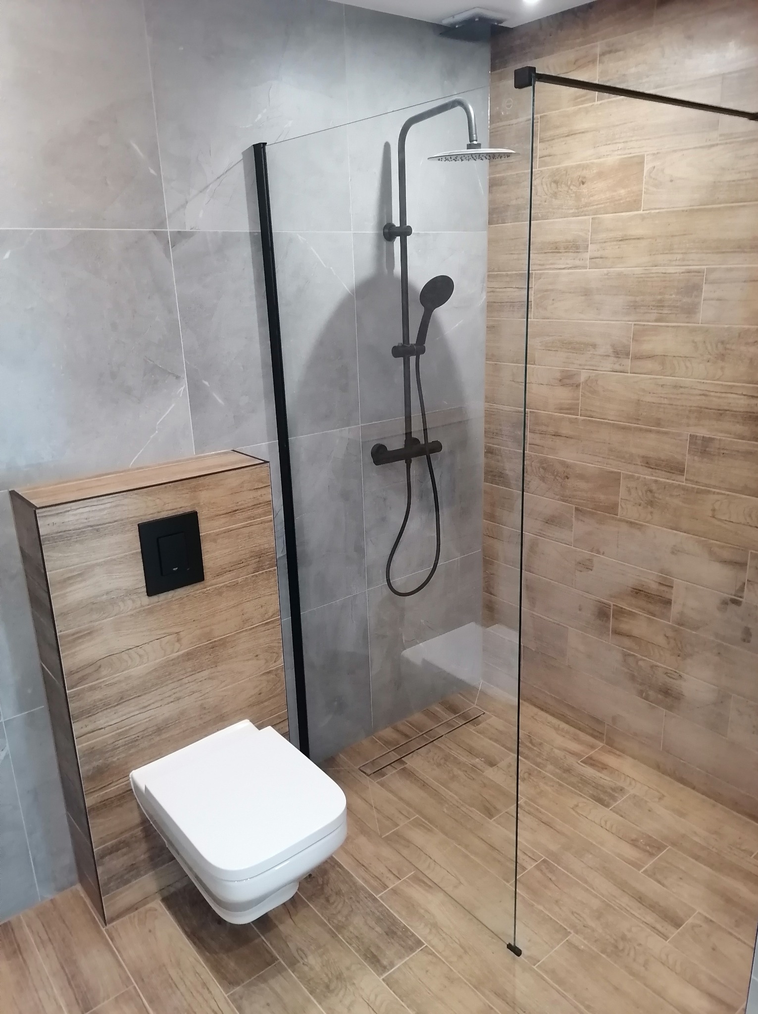 Nowoczesna kabina prysznicowa typu walk-in z czarnymi elementami armatury i odpływem liniowym, obok podwieszana toaleta z deską wolnoopadającą na tle płytek imitujących drewno i szary marmur.