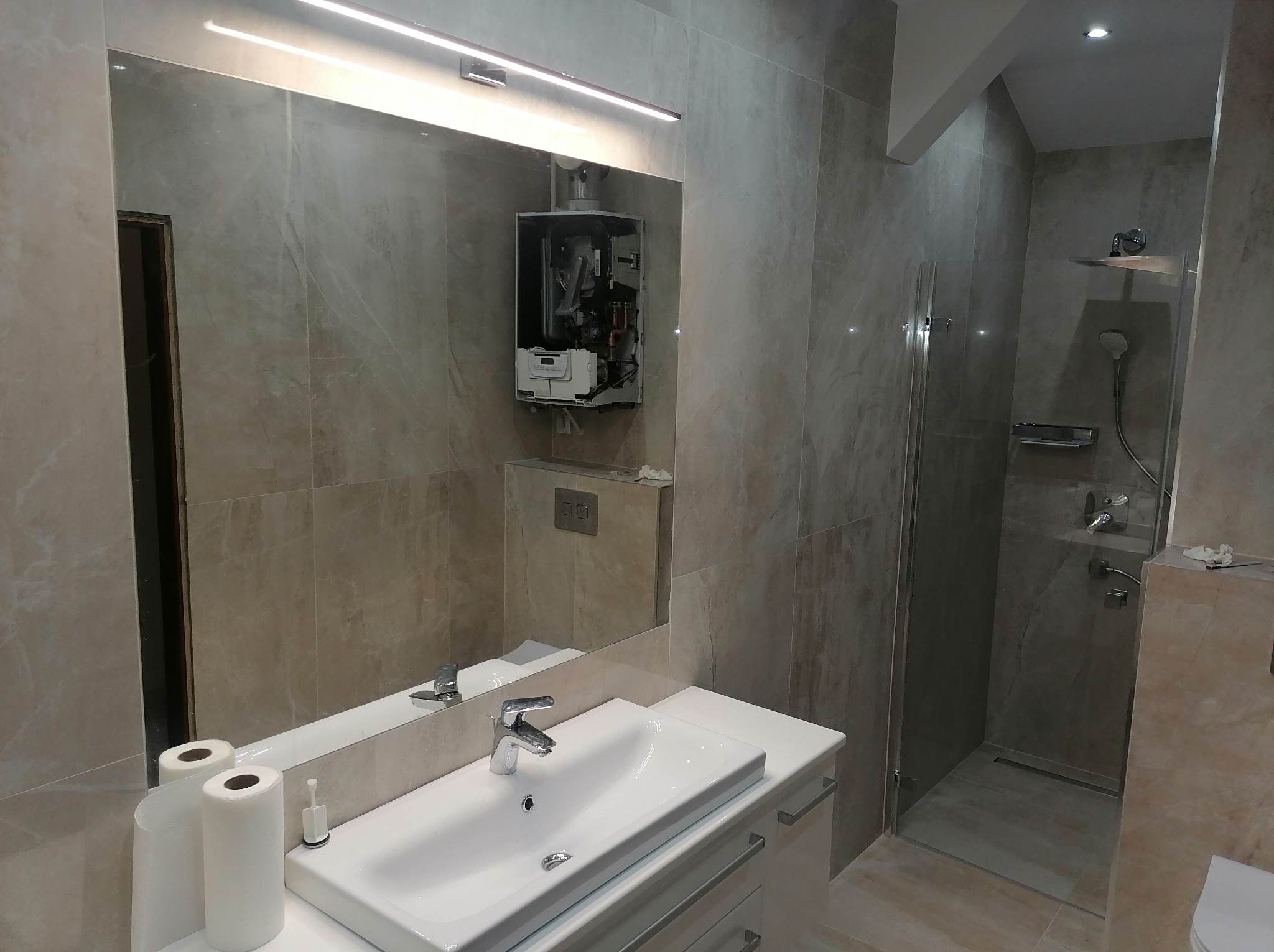 Wykończona łazienka z beżowymi płytkami imitującymi marmur, białą umywalką z szafką i kabiną prysznicową. W lustrze widoczny zamontowany, ale otwarty piec gazowy.