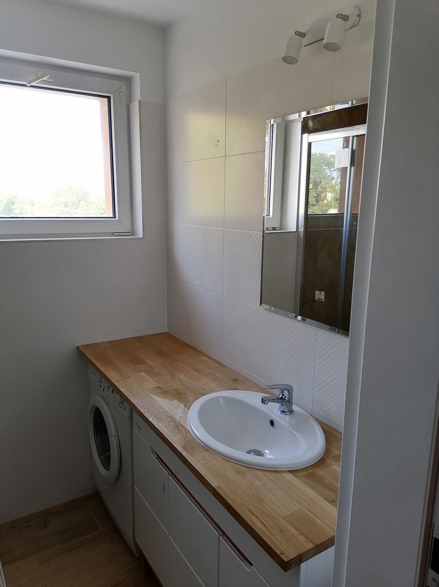 Nowoczesna łazienka z drewnianym blatem, białą umywalką wpuszczaną, pralką schowaną pod blatem, białą mozaiką na ścianie oraz lustrem z widokiem na zieleń za oknem.