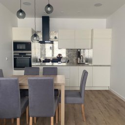 MZ Projektpodklucz - Jasna kuchnia z białymi szafkami i lustrzanym panelem nad blatem, widoczny stół z szarymi krzesłami i lampy wiszące.