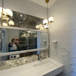 MZ Projektpodklucz - Wykończona łazienka z lustrem w ozdobnej ramie, umywalką z białego konglomeratu, dwoma suszarkami na ścianie i nowoczesnym oświetleniem.