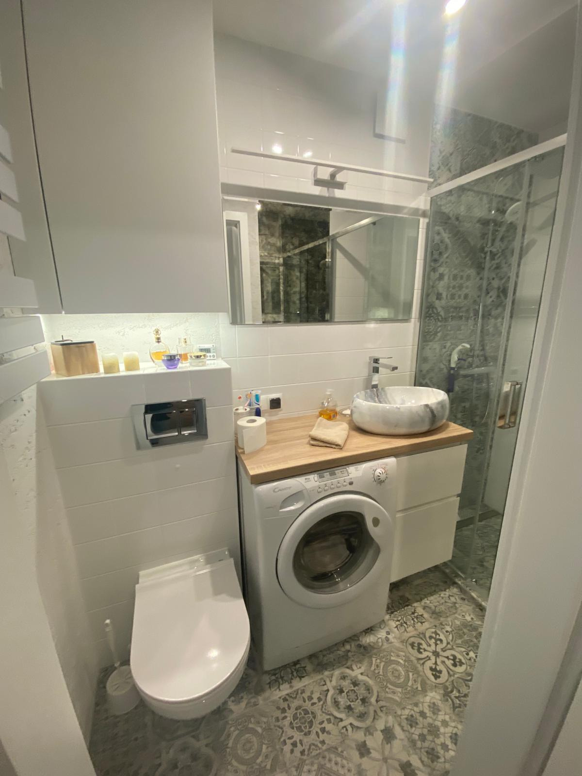 Nowoczesna łazienka z marmurową umywalką nablatową, białą pralką pod blatem, podwieszaną toaletą i kabiną prysznicową z dekoracyjnymi płytkami.