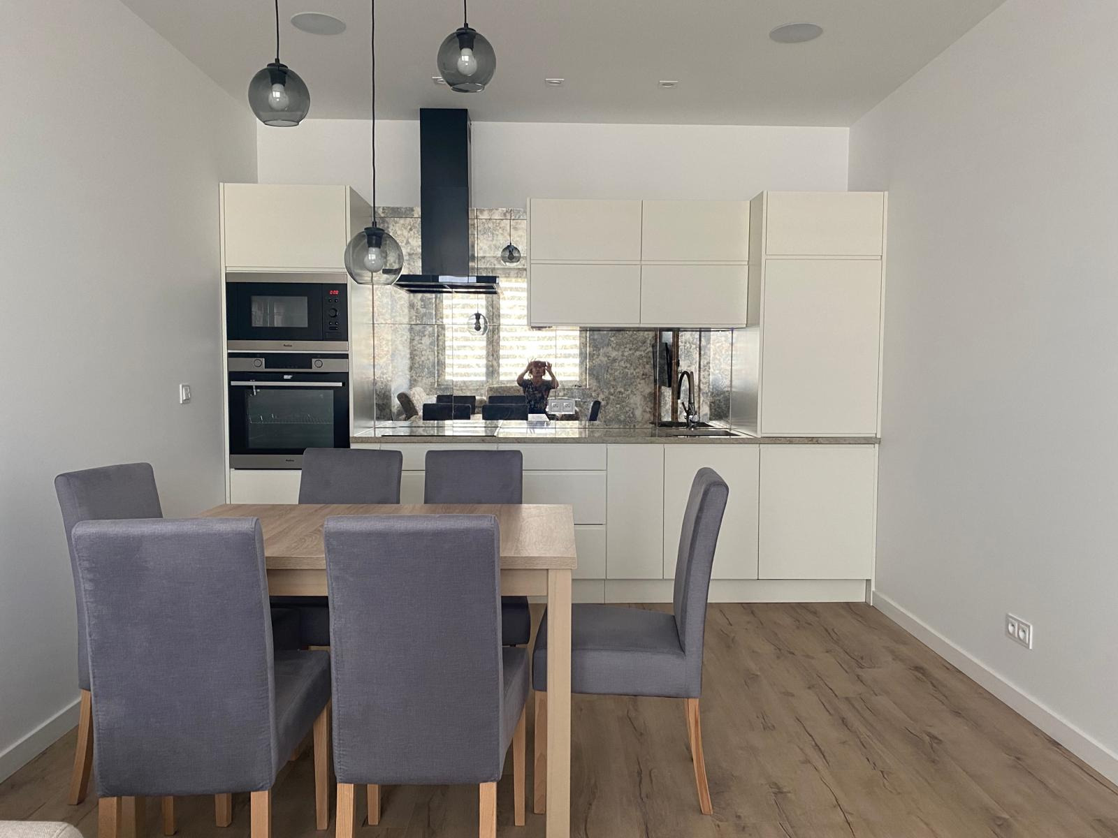 Jasna kuchnia z białymi szafkami i lustrzanym panelem nad blatem, widoczny stół z szarymi krzesłami i lampy wiszące.