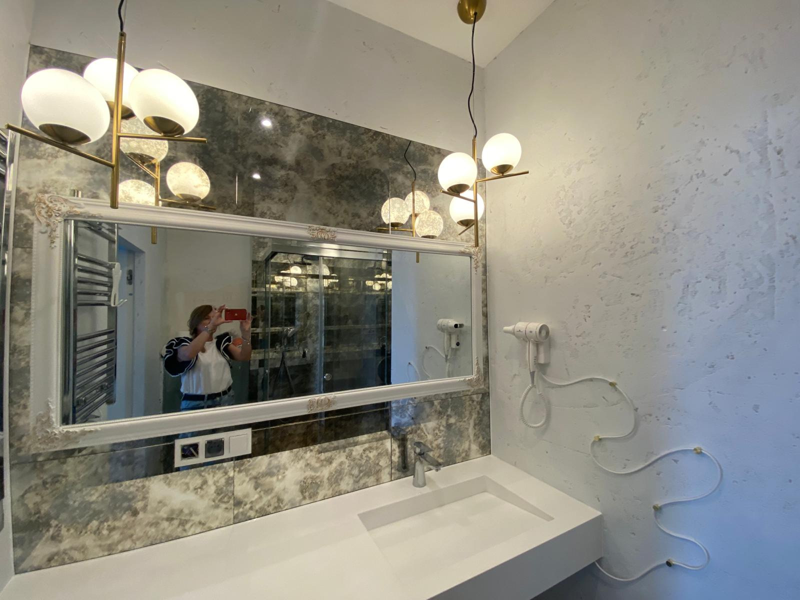 Wykończona łazienka z lustrem w ozdobnej ramie, umywalką z białego konglomeratu, dwoma suszarkami na ścianie i nowoczesnym oświetleniem.