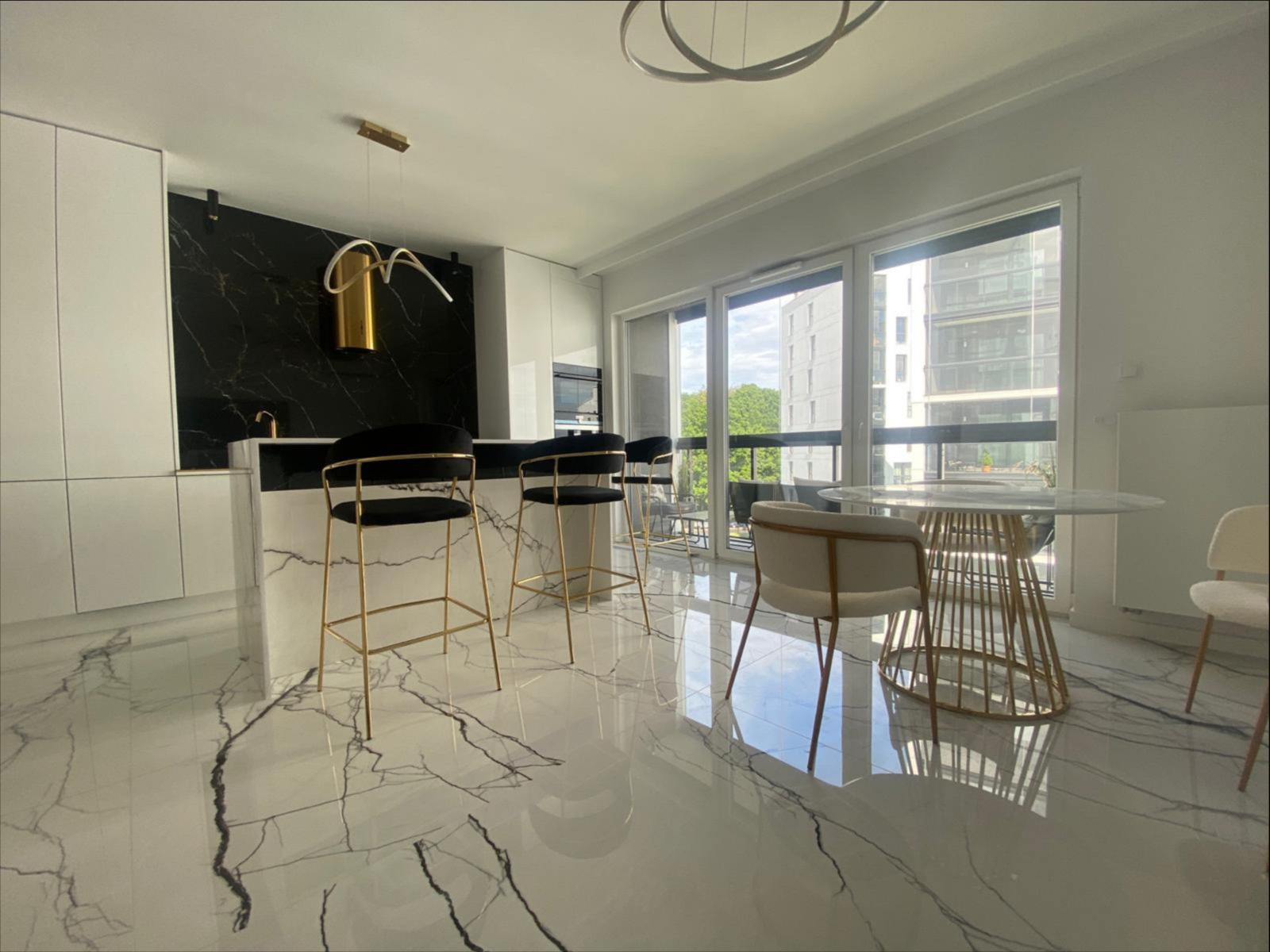 Nowoczesna kuchnia z wyspą, czarnym marmurem na ścianie, złotymi detalami oświetlenia i krzesłami barowymi, oraz marmurową posadzką z wyraźnym żyłowaniem, widok na zewnątrz przez duże okna.