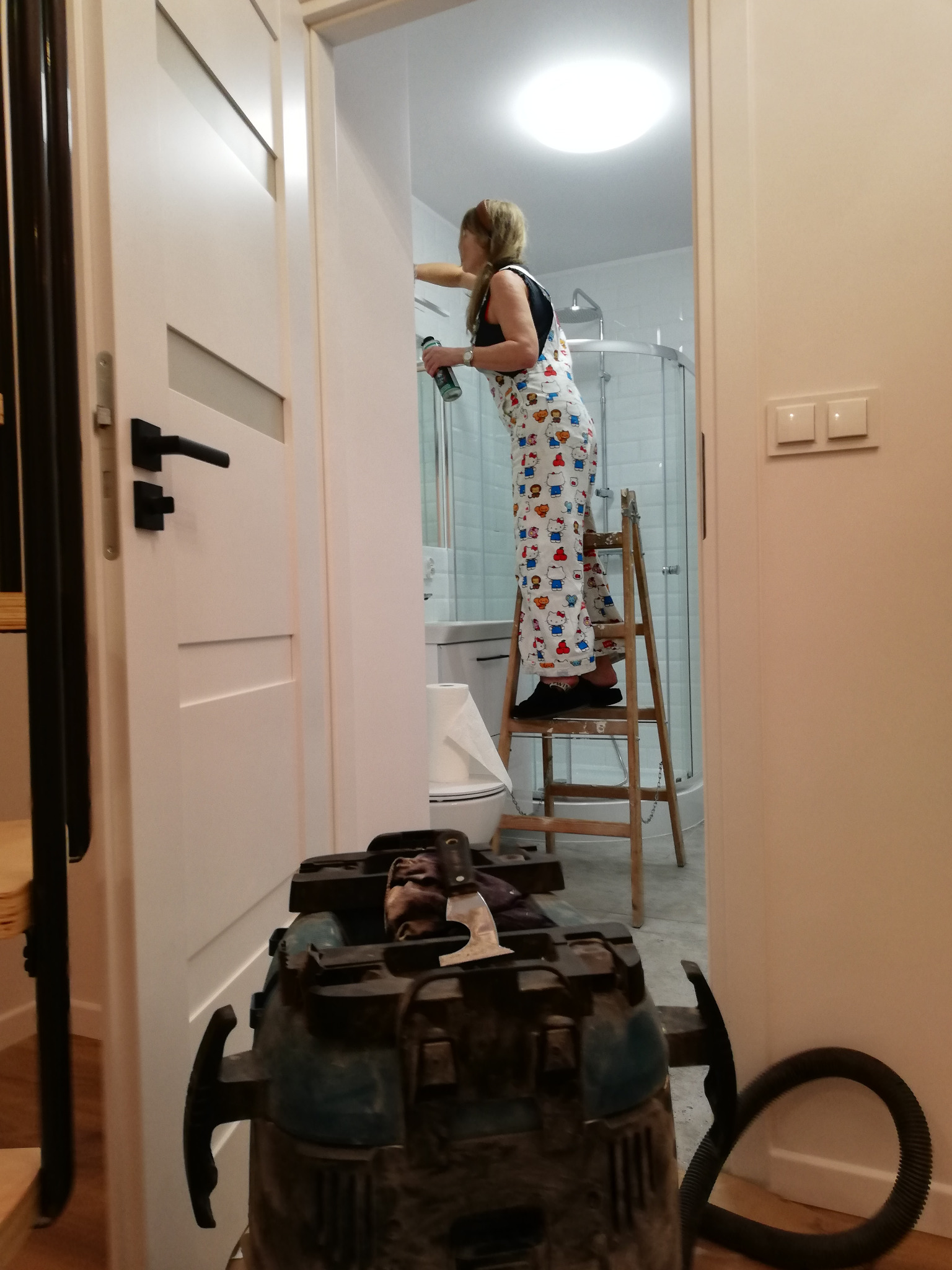 Kobieta w kombinezonie w Hello Kitty stoi na drewnianej drabinie i maluje ścianę w łazience z kabiną prysznicową; w tle widoczny odkurzacz przemysłowy.