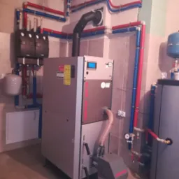 Kotłownia z nowoczesnym piecem DEFRO, zbiornikiem akumulacyjnym i instalacją grzewczą z rur w izolacji czerwono-niebieskiej, widoczna instalacja elektryczna oraz fragment ściany z płyt...