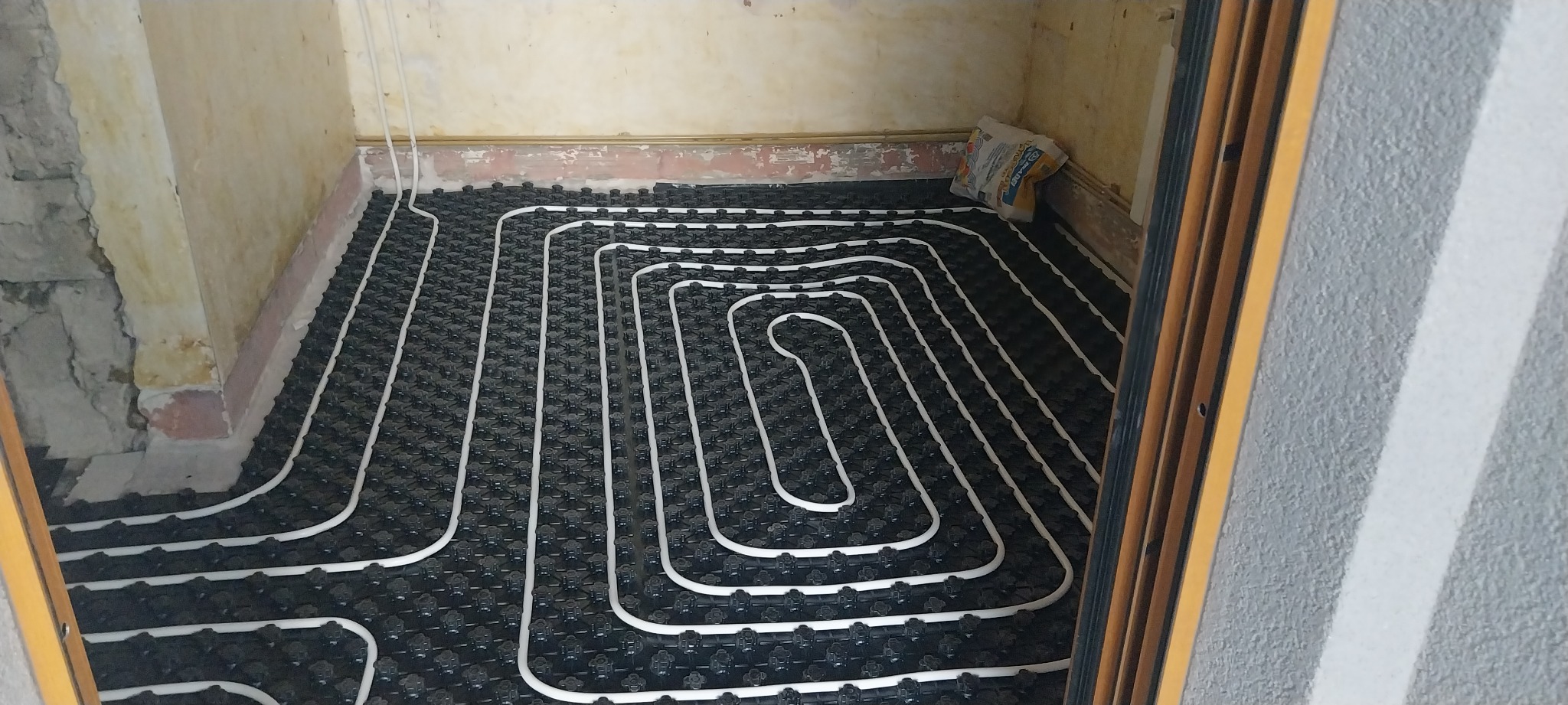 Instalacja ogrzewania podłogowego z białych rurek na czarnej macie w trakcie montażu w pomieszczeniu ze ścianami w stanie surowym.