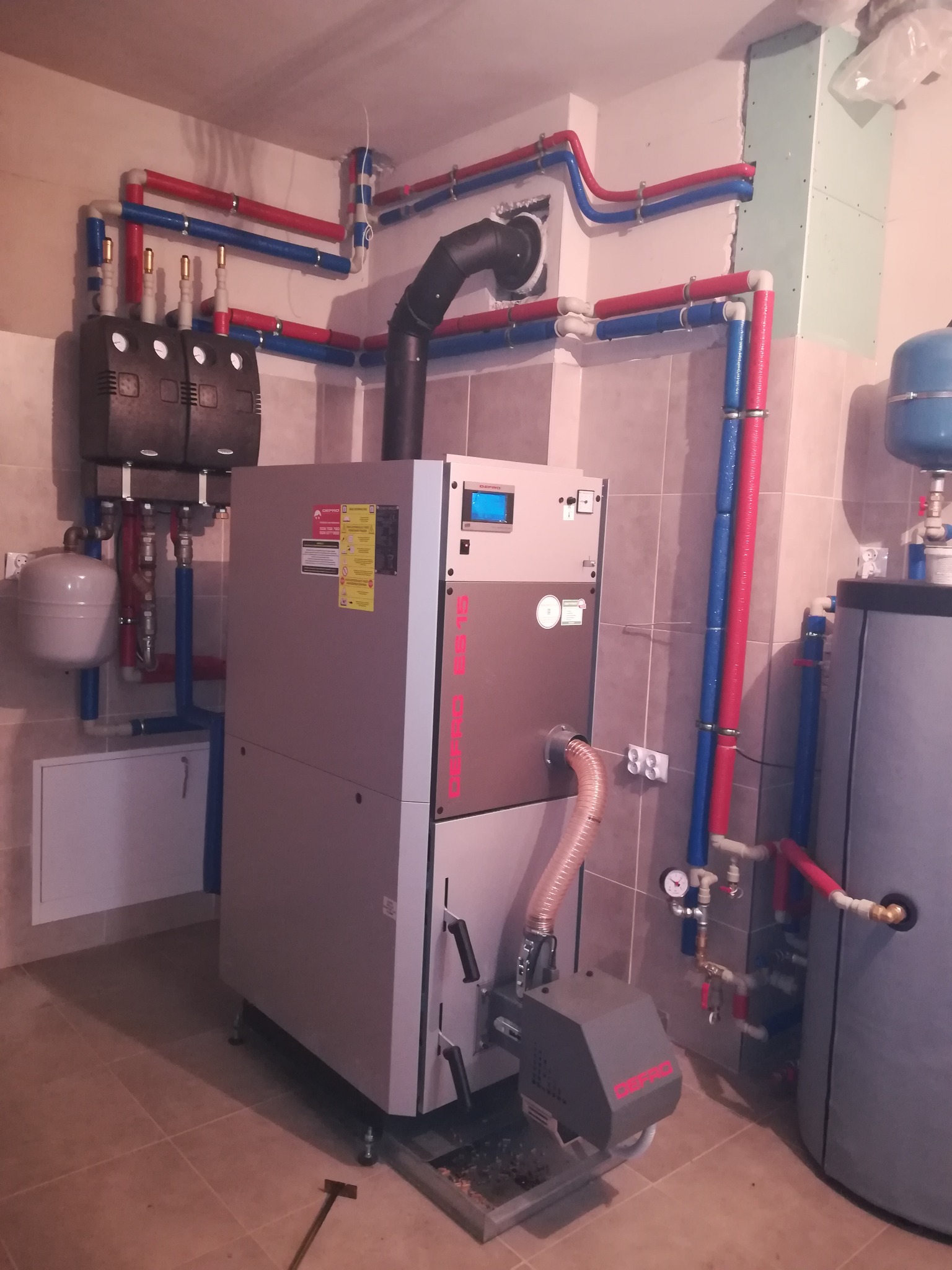 Kotłownia z nowoczesnym piecem DEFRO, zbiornikiem akumulacyjnym i instalacją grzewczą z rur w izolacji czerwono-niebieskiej, widoczna instalacja elektryczna oraz fragment ściany z płyt...