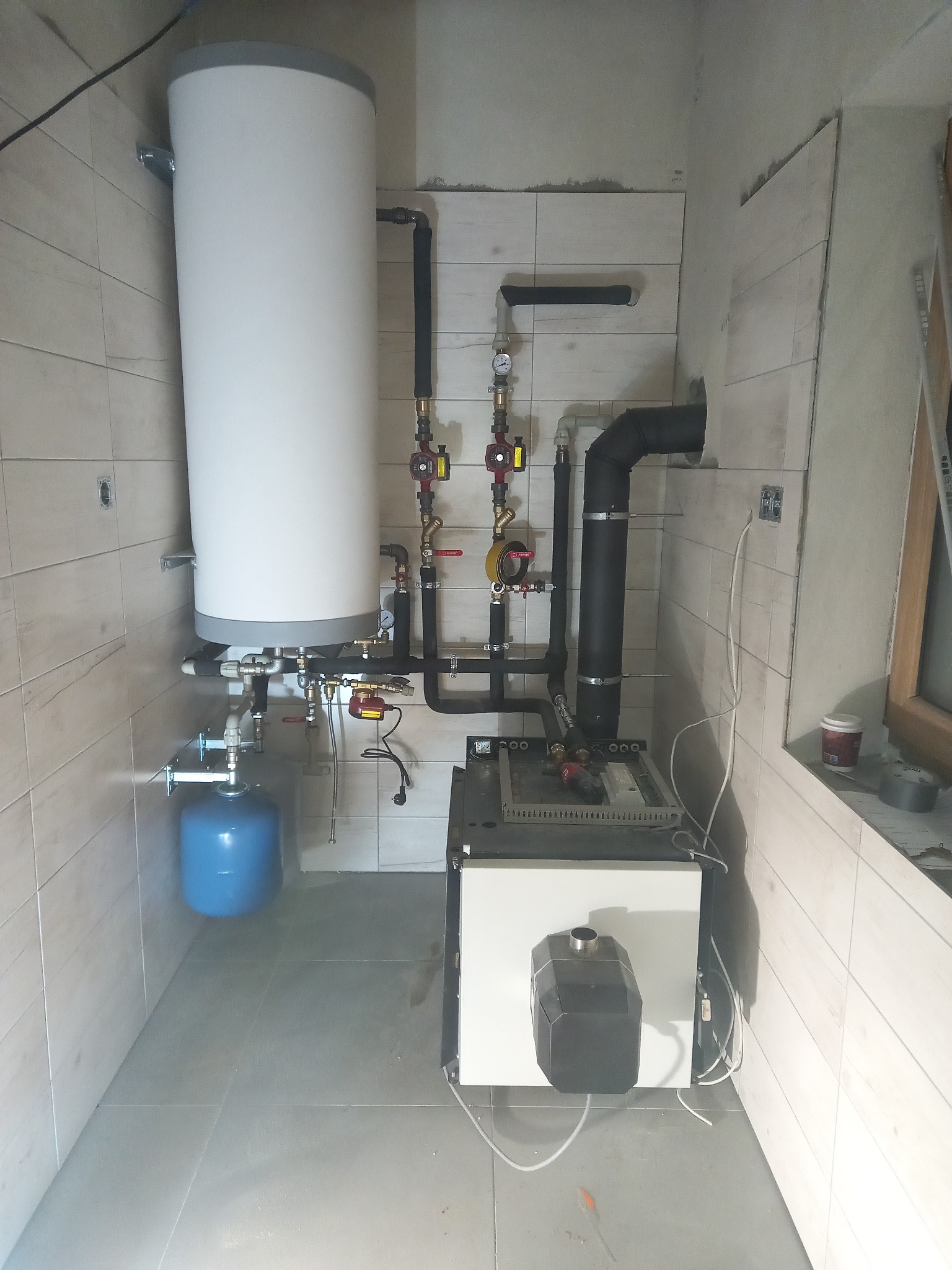 Nowa instalacja grzewcza w kotłowni z białym zasobnikiem ciepłej wody, niebieskim naczyniem przeponowym i czarnym kotłem grzewczym, rury w izolacji, płytki ceramiczne na ścianach w trakcie montażu.
