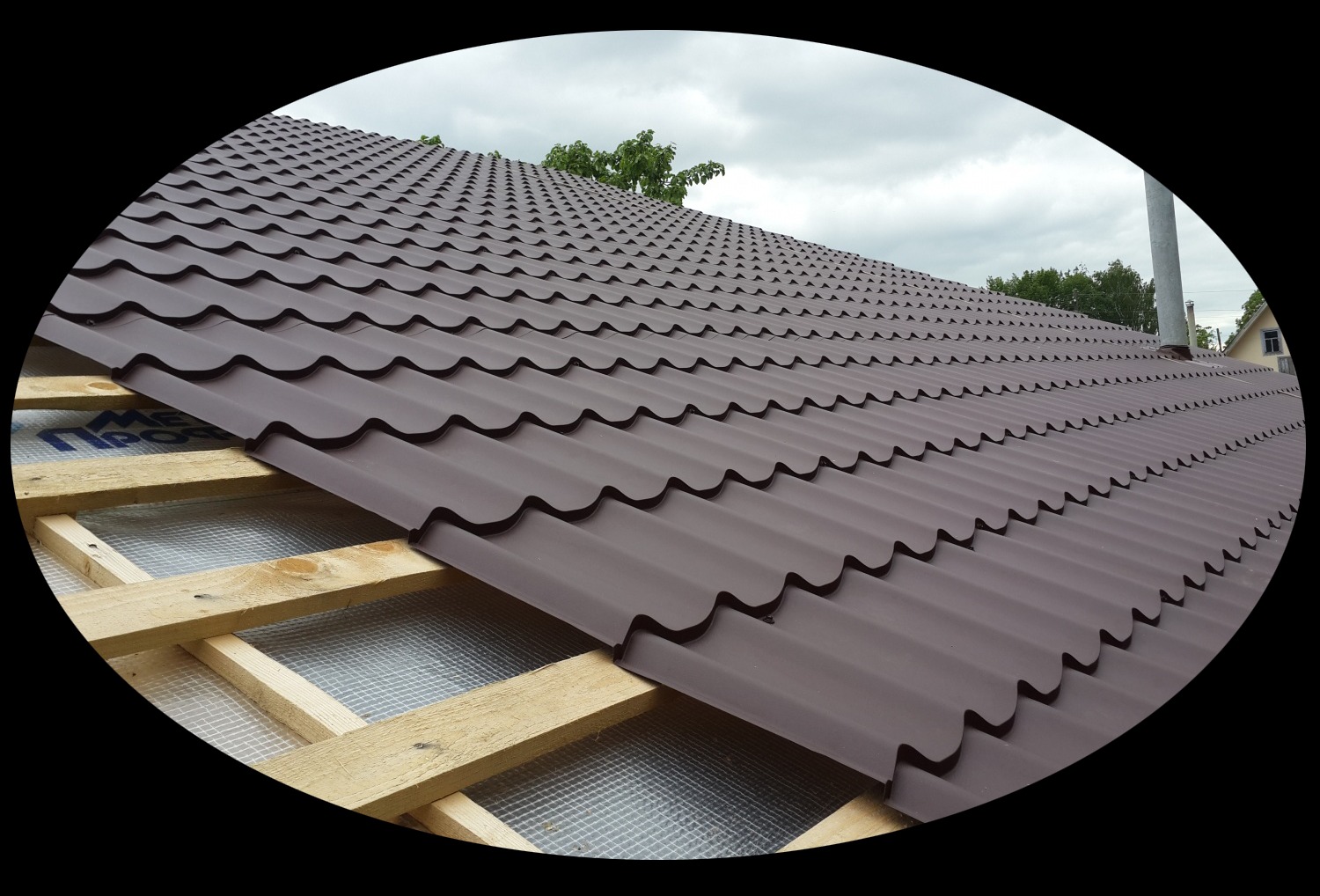 Widok na częściowo pokryty dach z brązowej blachodachówki, widoczna konstrukcja drewniana i warstwa izolacyjna, na tle pochmurnego nieba.