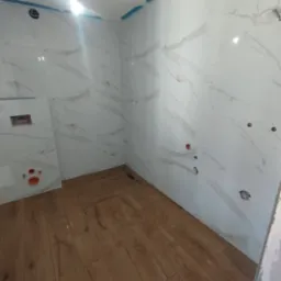 Wnętrze łazienki w trakcie prac wykończeniowych: białe płytki imitujące marmur na ścianach z widocznymi otworami na instalacje, drewnopodobne panele na podłodze, fragment odpływu liniowego.