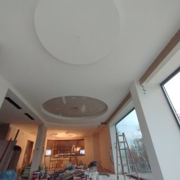 Usługi remontowe Patryk Nowacki - Wnętrze w trakcie prac wykończeniowych: biały sufit z okrągłymi elementami dekoracyjnymi, widoczne narzędzia, drabina oparta o duże okno, ślady po pracach na ścianach.