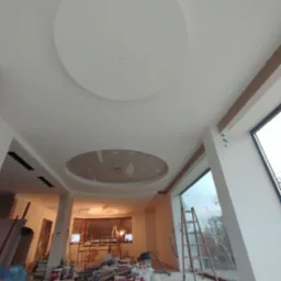 Wnętrze w trakcie prac wykończeniowych: biały sufit z okrągłymi elementami dekoracyjnymi, widoczne narzędzia, drabina oparta o duże okno, ślady po pracach na ścianach.