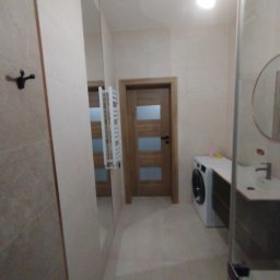 Usługi remontowe Patryk Nowacki - Jasna łazienka z beżowymi płytkami, drewnianymi drzwiami z przeszkleniami, pralką i umywalką z szafką, widok z kabiny prysznicowej.