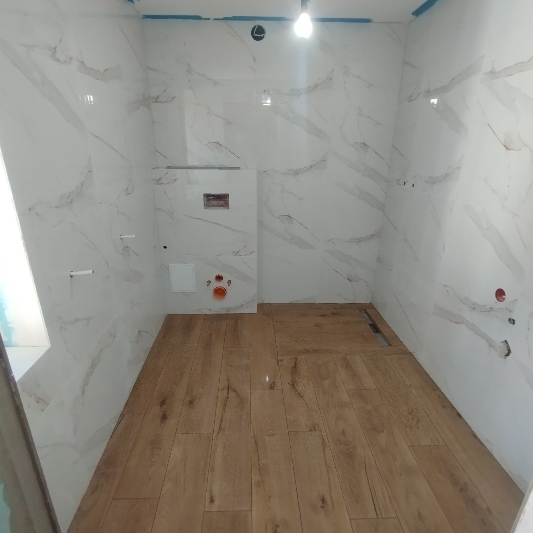 Wnętrze łazienki w Kaliszu w trakcie remontu: białe, marmurkowe płytki na ścianach kontrastują z panelami podłogowymi imitującymi drewno, widoczne instalacje hydrauliczne i otwory na armaturę.