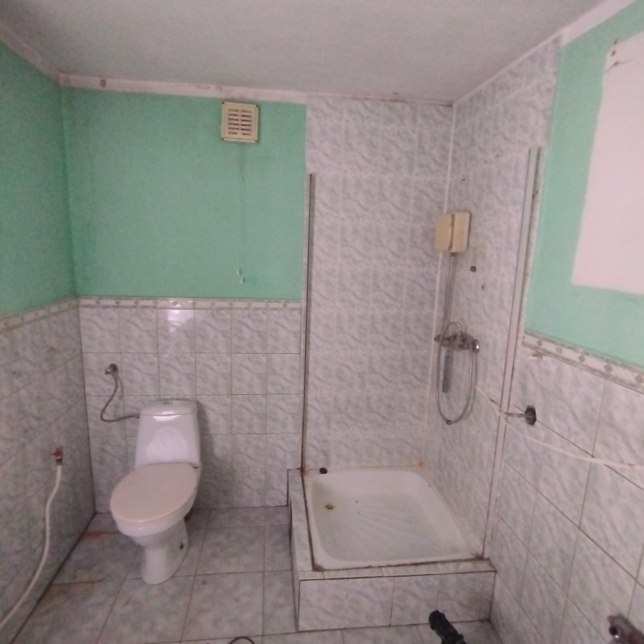 Łazienka z toaletą w kolorze różowym i brodzikiem, wykończona płytkami w odcieniach szarości, z widoczną instalacją hydrauliczną i fragmentem zielonej ściany.