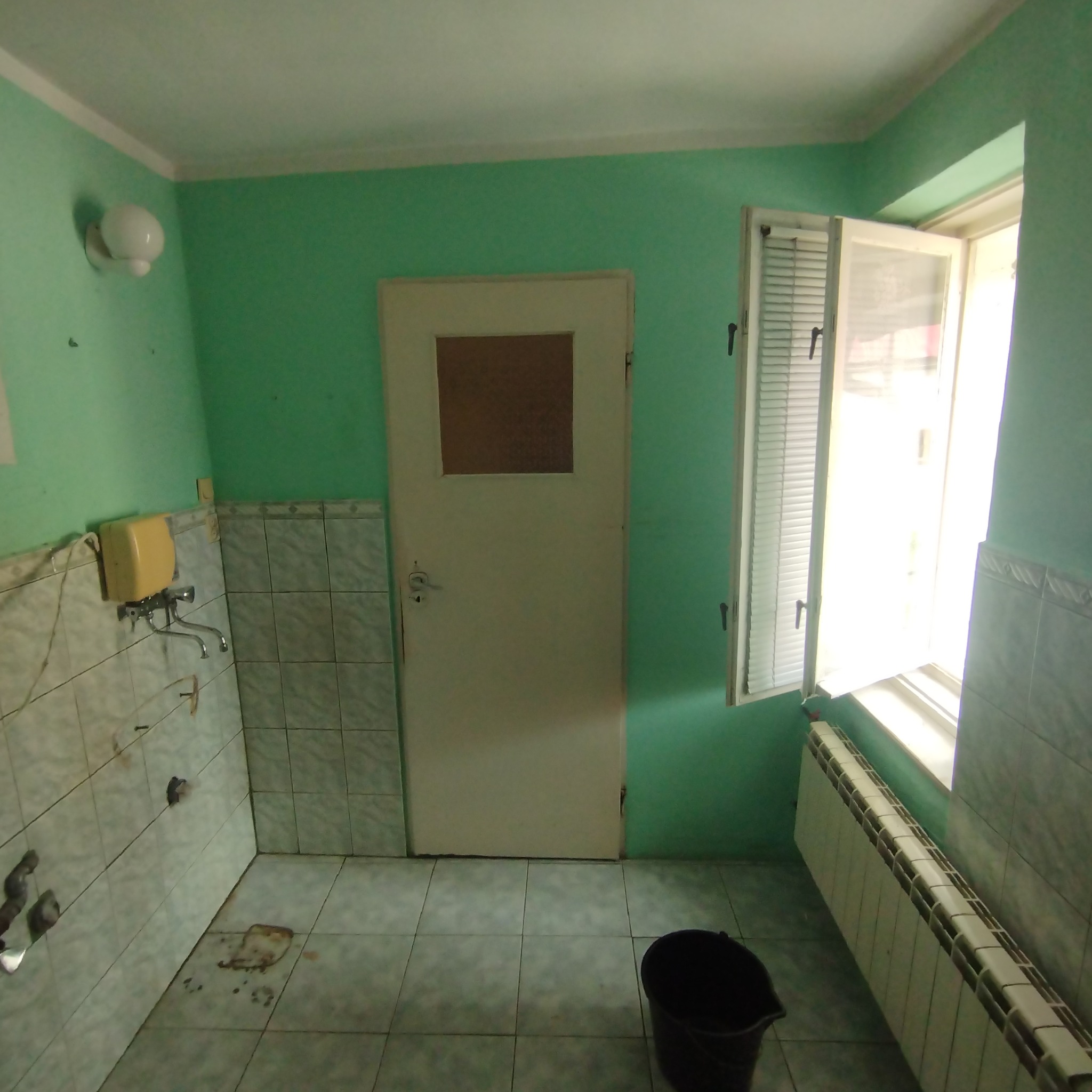 Łazienka w trakcie remontu: zielone ściany, stare płytki, otwarte okno z żaluzjami, widoczny grzejnik i wiadro na podłodze.