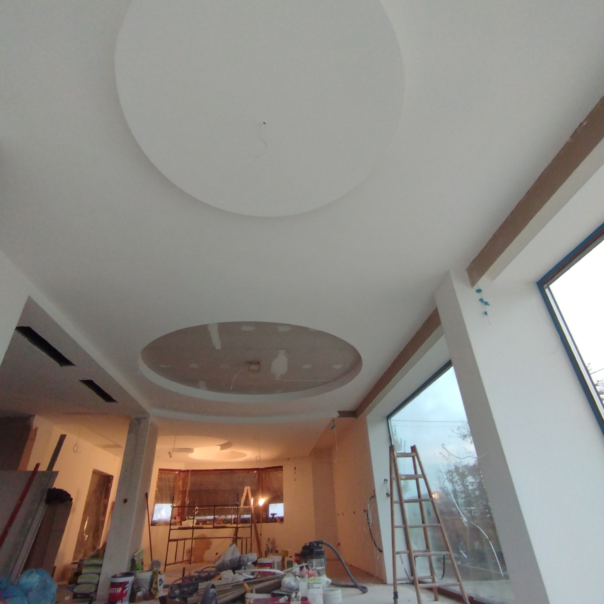 Wnętrze w trakcie prac wykończeniowych: biały sufit z okrągłymi elementami dekoracyjnymi, widoczne narzędzia, drabina oparta o duże okno, ślady po pracach na ścianach.