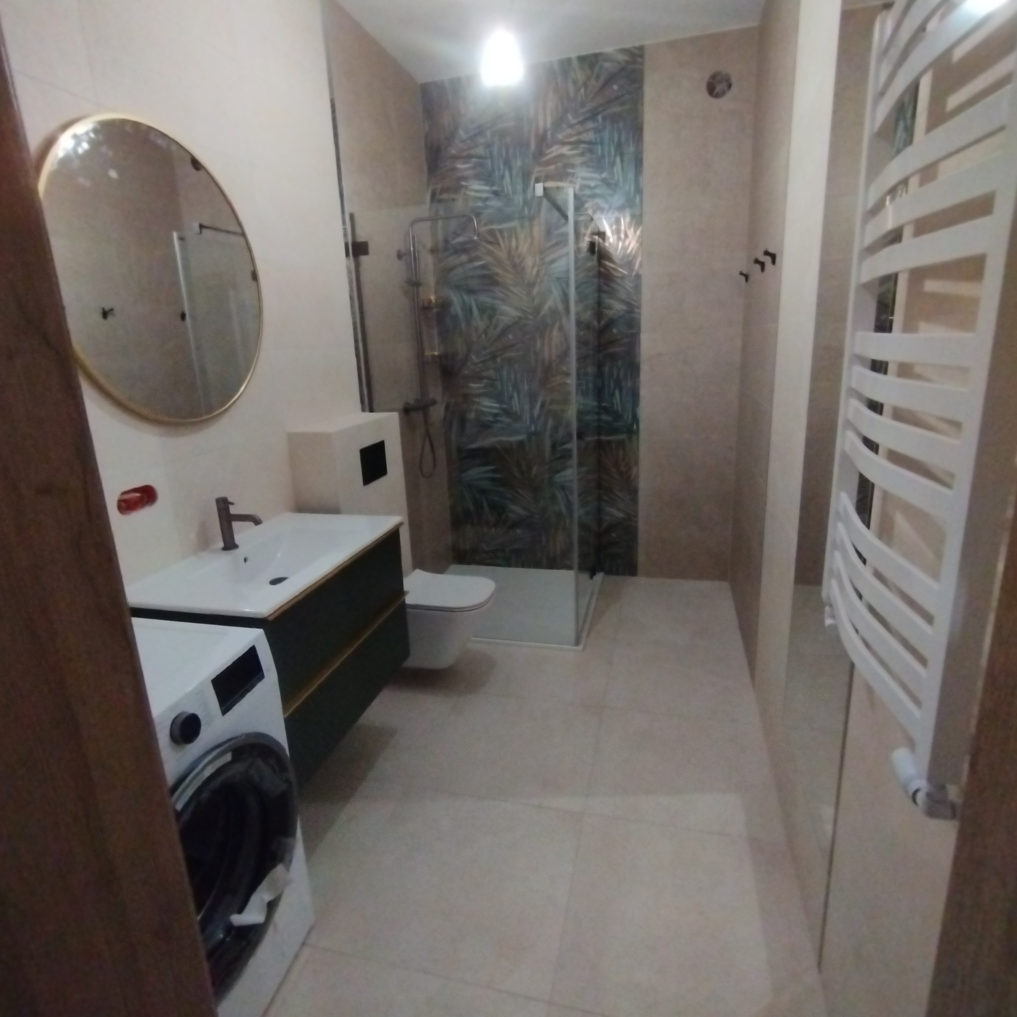 Nowoczesna łazienka z kabiną prysznicową typu walk-in, umywalką z szafką w kolorze ciemnej zieleni, okrągłym lustrem w złotej ramie i grzejnikiem drabinkowym.