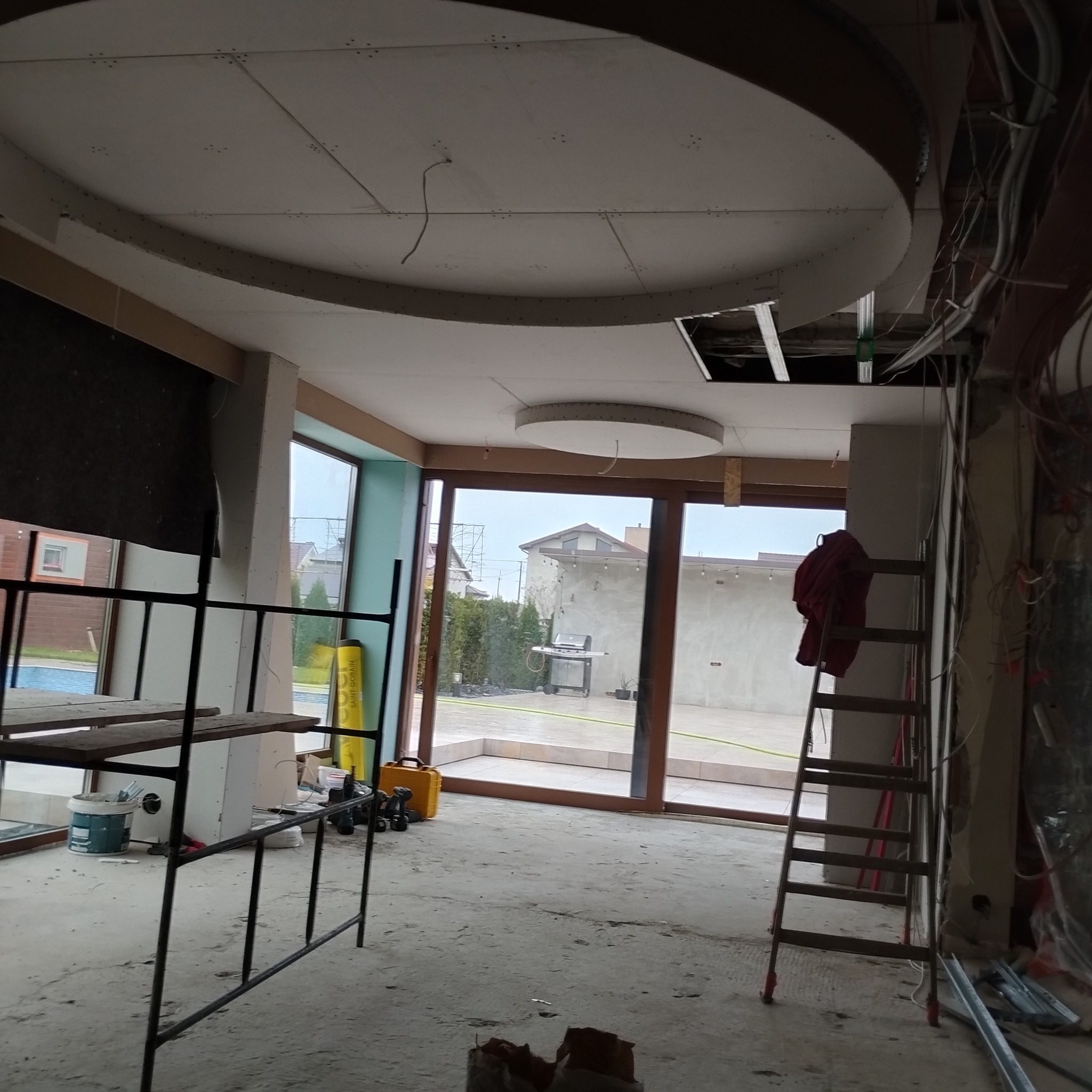 Wnętrze w trakcie prac wykończeniowych: sufit z elementami gipsowo-kartonowymi o nietypowym kształcie, widoczne rusztowanie, drabina i okna z widokiem na ogród.