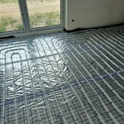 Białe rurki ogrzewania podłogowego marki Keller System ułożone na folii aluminiowej z czarną siatką w nowym budynku, widoczne okno Salamander