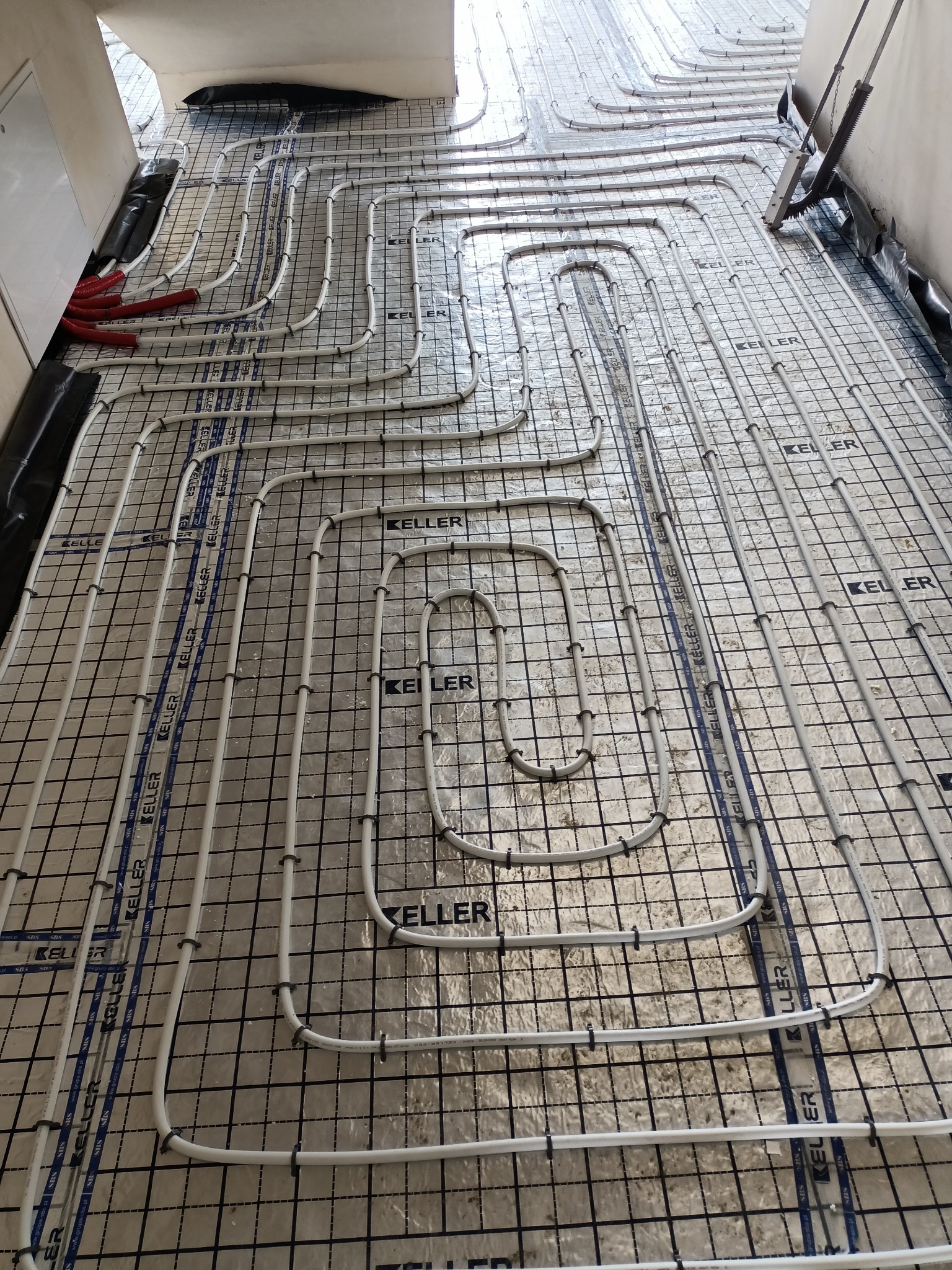 Białe rury ogrzewania podłogowego ułożone na srebrnej folii z czarną siatką, widoczne oznaczenia producenta Keller na folii.