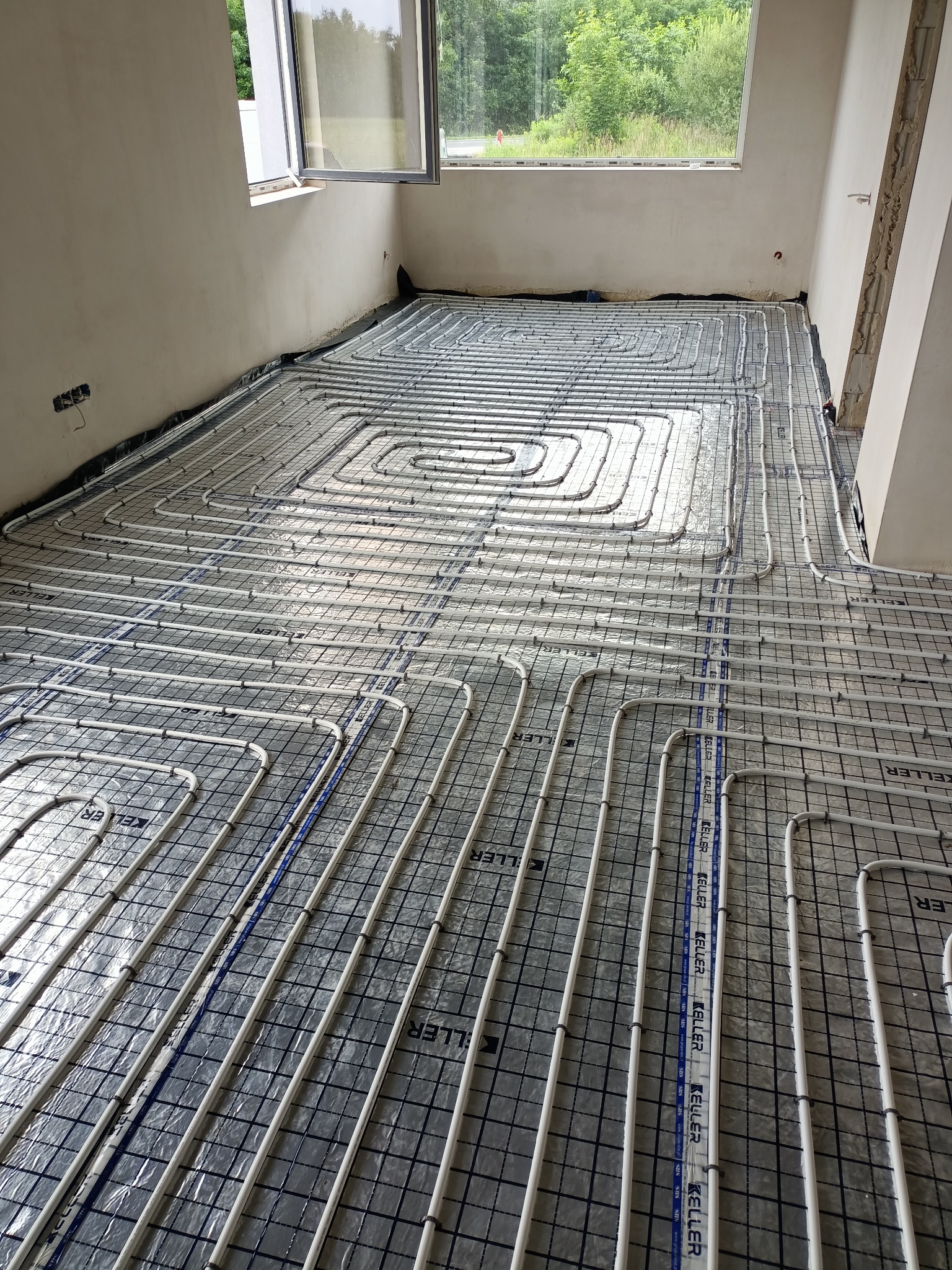 Białe rury ogrzewania podłogowego ułożone w pętle na srebrnej folii z rastrem, widok z góry w surowym wnętrzu z oknem.