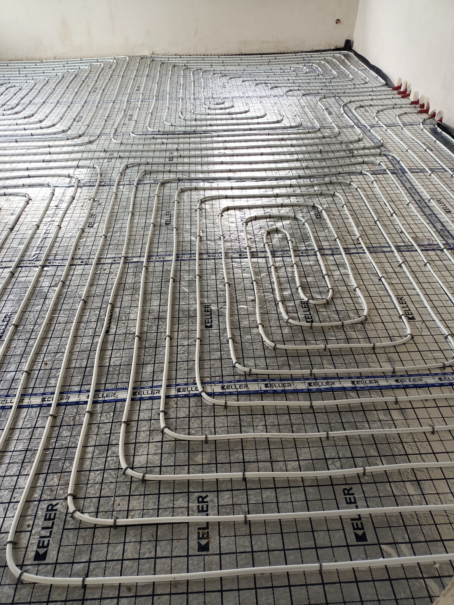 Białe rury ogrzewania podłogowego ułożone w pętle na srebrnej folii izolacyjnej, przymocowane do czarnej siatki z widocznym logo 'KELLER', przygotowane do zalania wylewką.