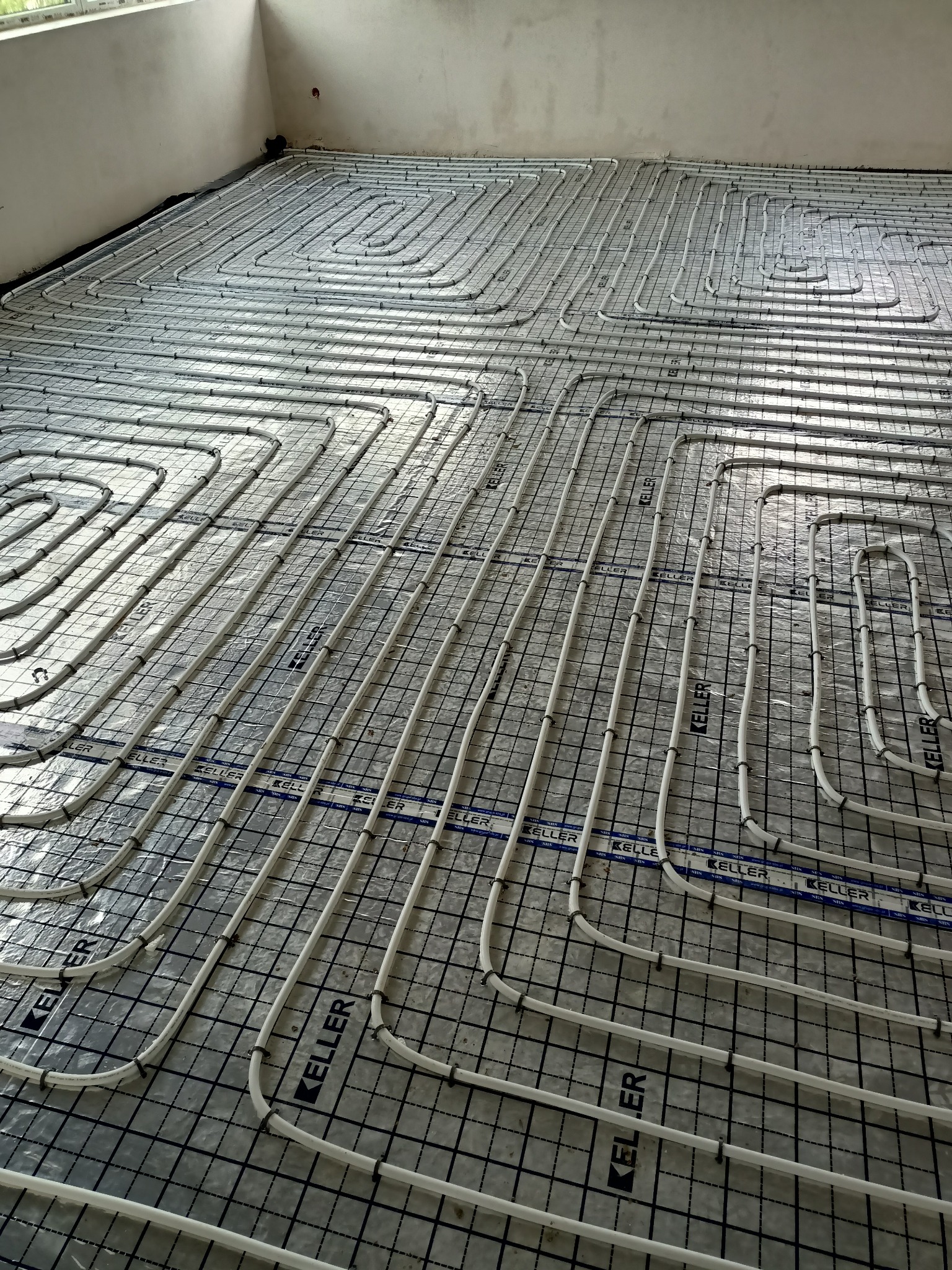 Białe rury ogrzewania podłogowego ułożone spiralnie na siatce z tworzywa sztucznego, widoczne mocowania rur i folia izolacyjna pod siatką, surowy stan wykończenia pomieszczenia.