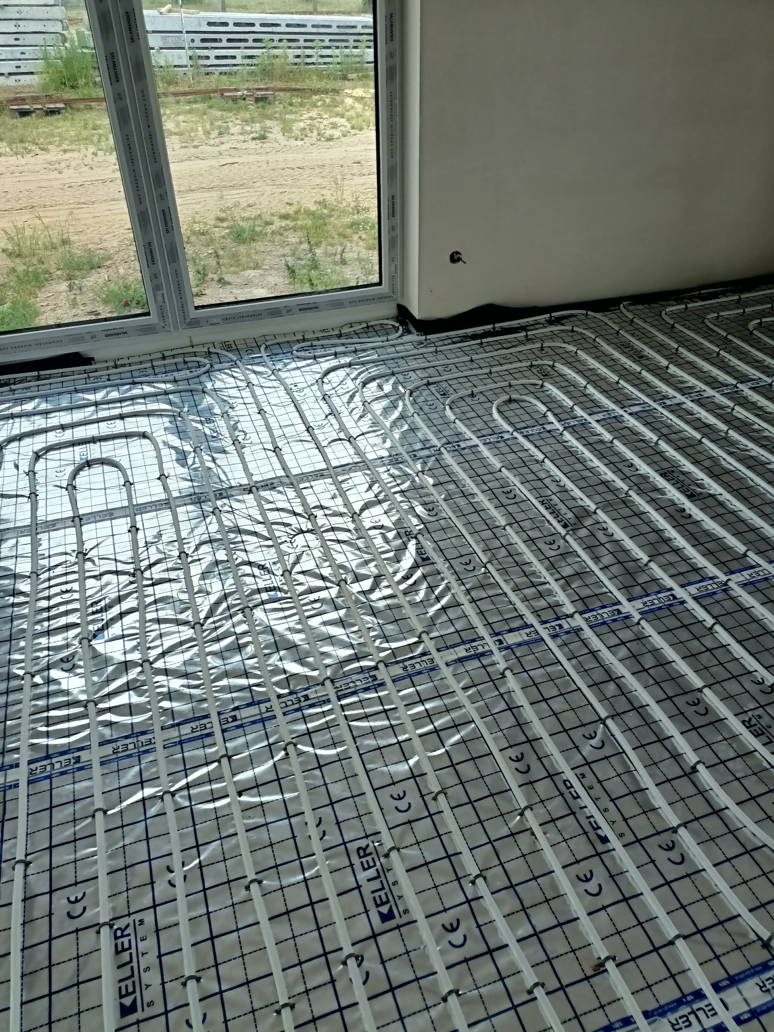 Białe rurki ogrzewania podłogowego marki Keller System ułożone na folii aluminiowej z czarną siatką w nowym budynku, widoczne okno Salamander