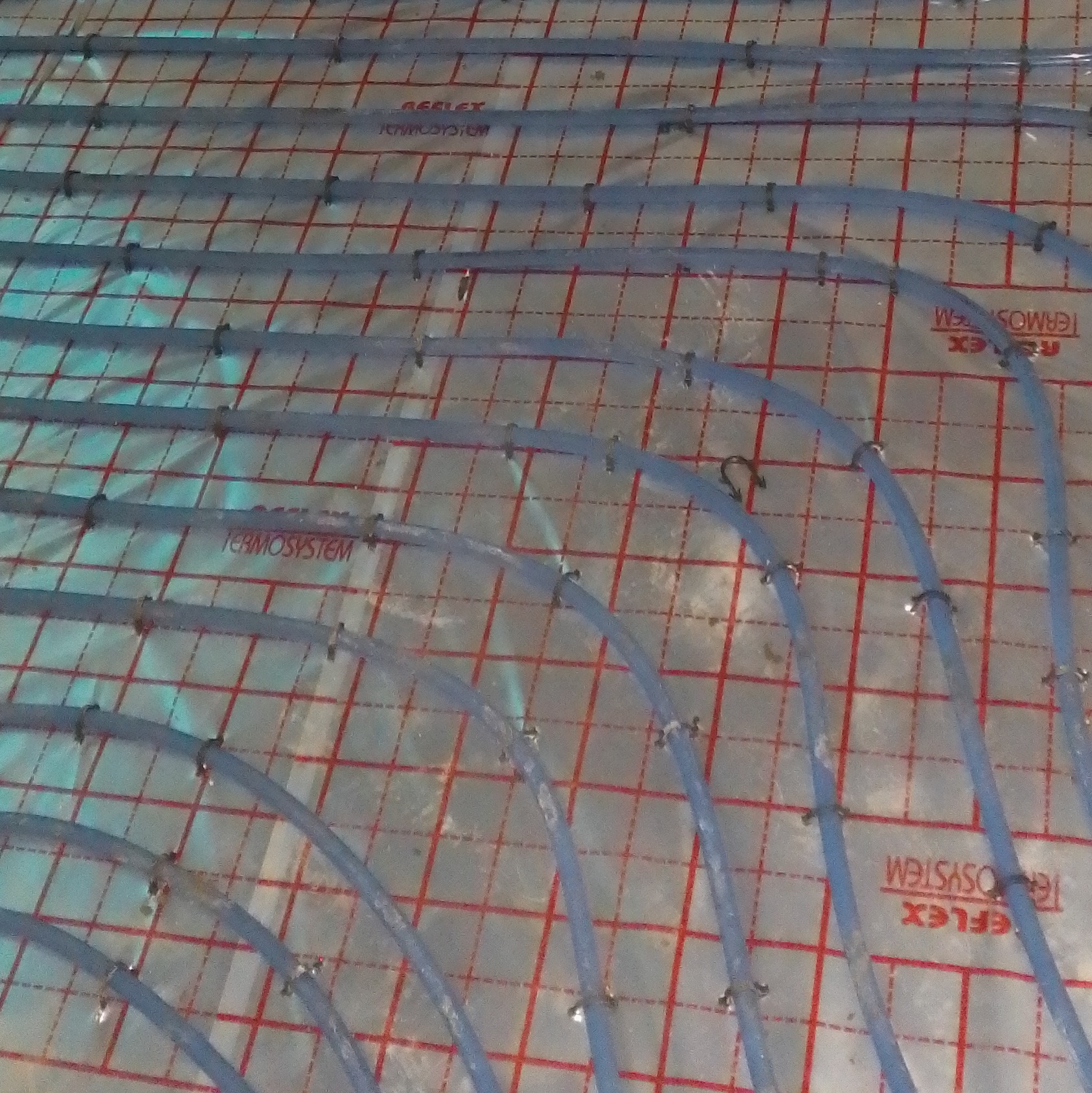 Niebieskie rury ogrzewania podłogowego, przymocowane metalowymi klamrami do folii z czerwoną siatką, widok z góry na etapie instalacji.