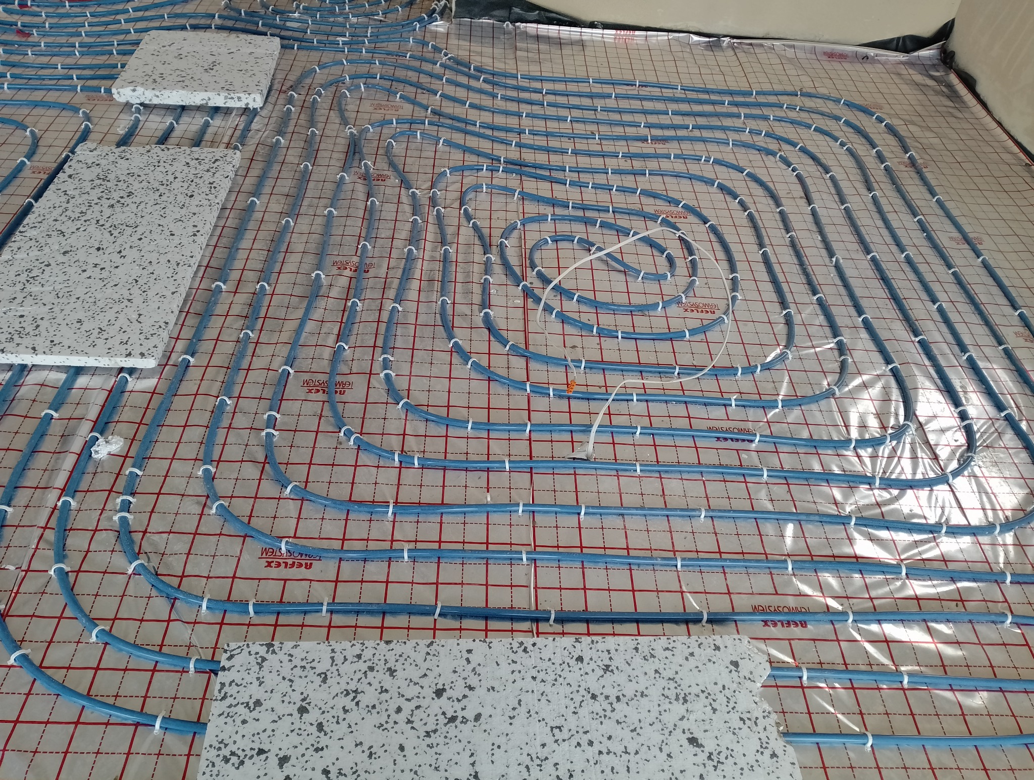 Rozłożone niebieskie rury systemu grzewczego na folii z siatką, częściowo przykryte szarymi płytkami z wzorem lastryko.