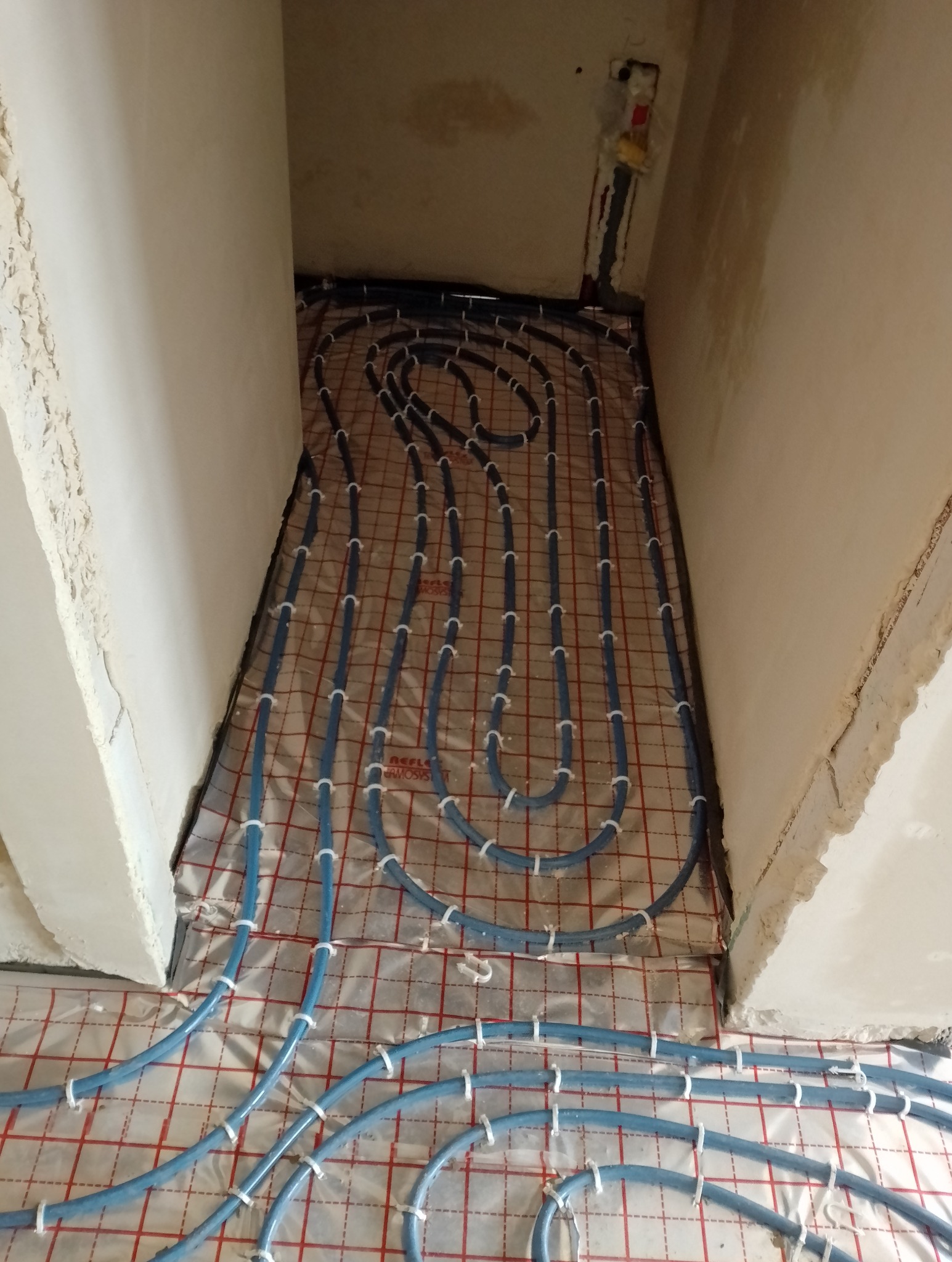 Niebieskie rury instalacji grzewczej, mocowane białymi uchwytami, rozłożone na folii z czerwoną siatką, widok na surowy stan podłogi przed wylewką.