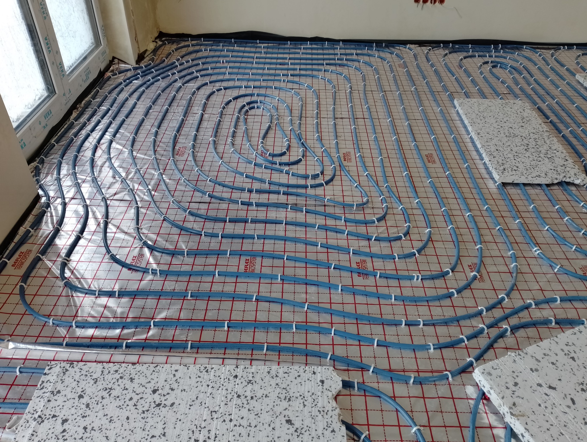 Rozłożone niebieskie rury systemu grzewczego na folii z siatką, częściowo przykryte białymi płytami izolacyjnymi, widok z góry.