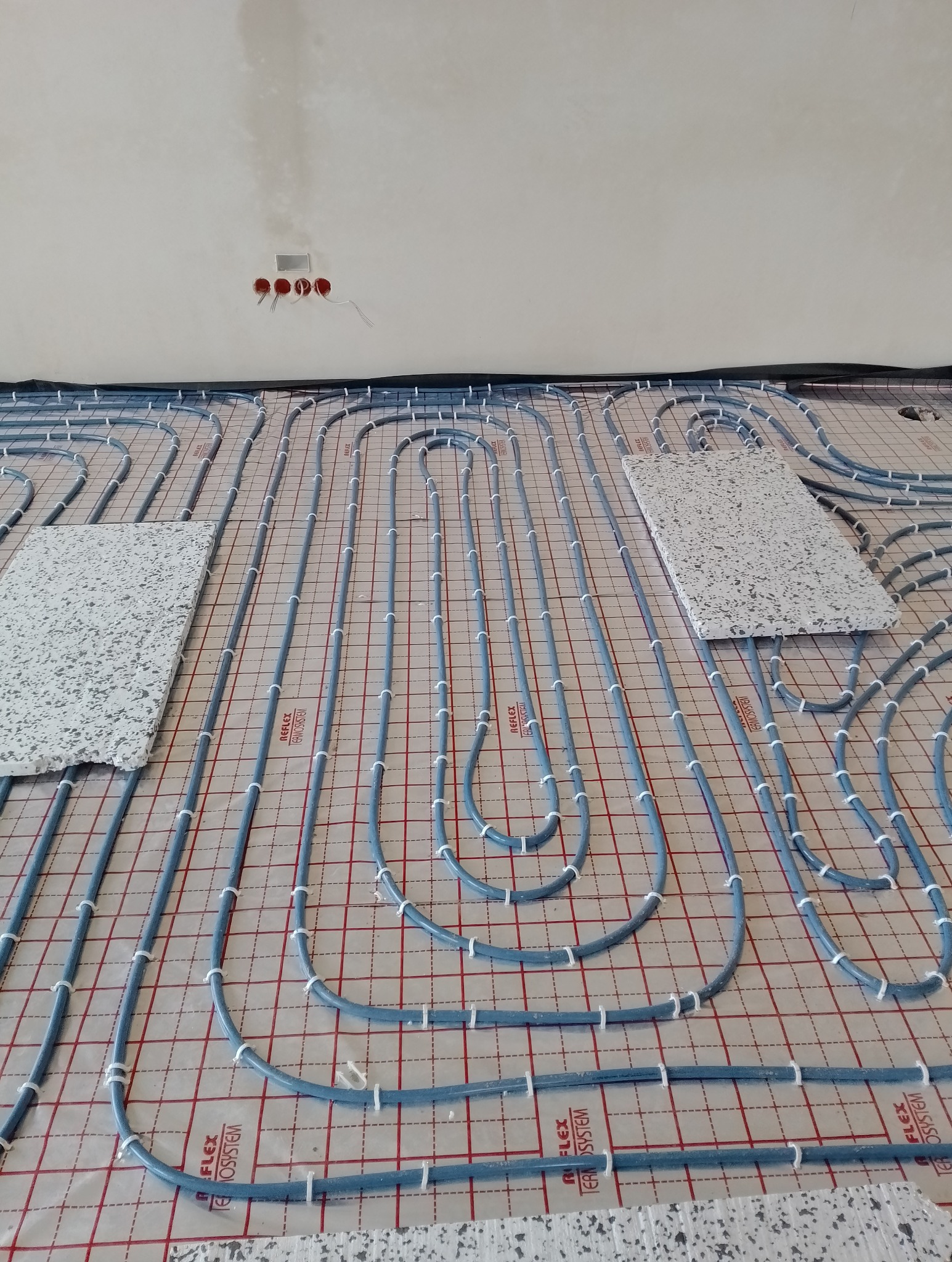 Rozłożone rury w systemie montażu na macie z rastrem, przygotowane do zalania wylewką, widoczne fragmenty izolacji brzegowej, na ścianie puszki elektryczne.