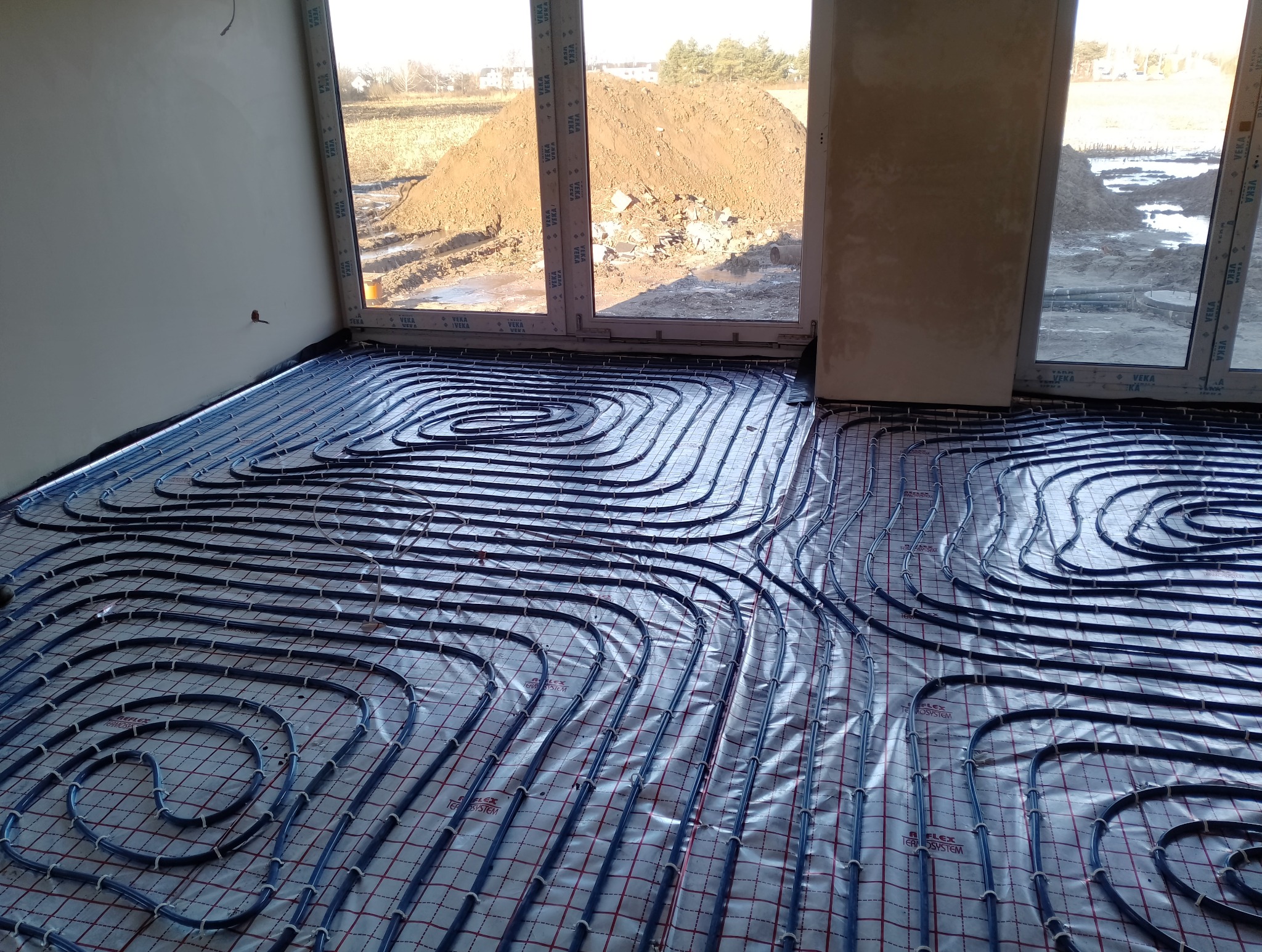 Rozłożone rury systemu grzewczego na siatce w surowym wnętrzu, widok przez okno na plac budowy z hałdą ziemi.