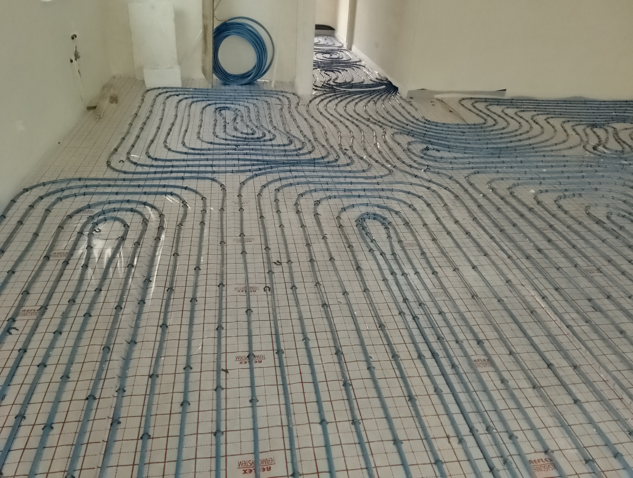 Rozłożone rury systemu grzewczego na podłodze z widoczną siatką montażową i zwiniętym niebieskim wężem obok ściany.