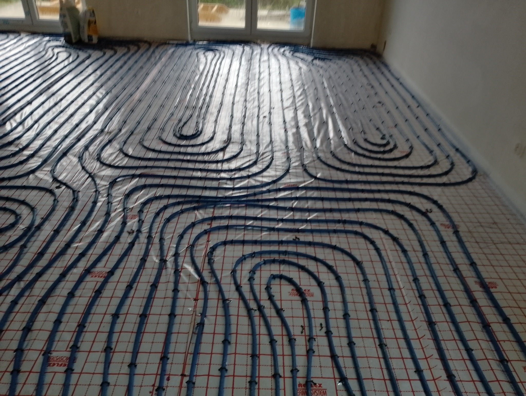 Rozłożone rury ogrzewania podłogowego na folii z siatką, widoczne złącza i fragmenty ścian pomieszczenia.