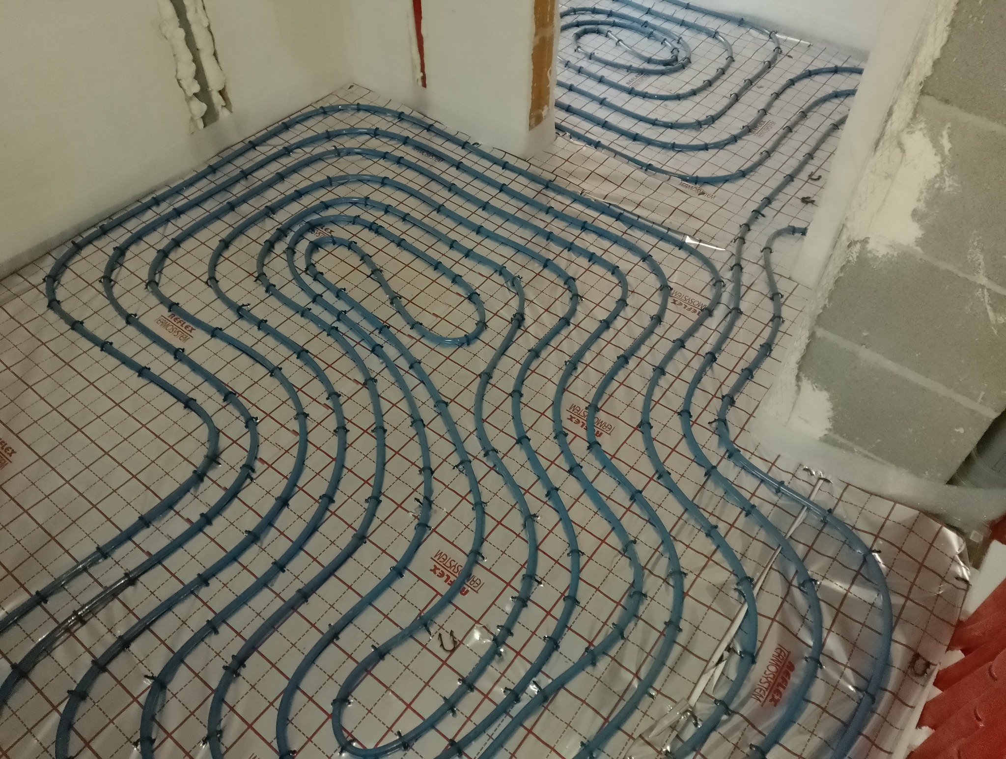Rozłożone niebieskie rury systemu ogrzewania podłogowego na izolacyjnej macie z nadrukowaną siatką w pomieszczeniu w trakcie instalacji.