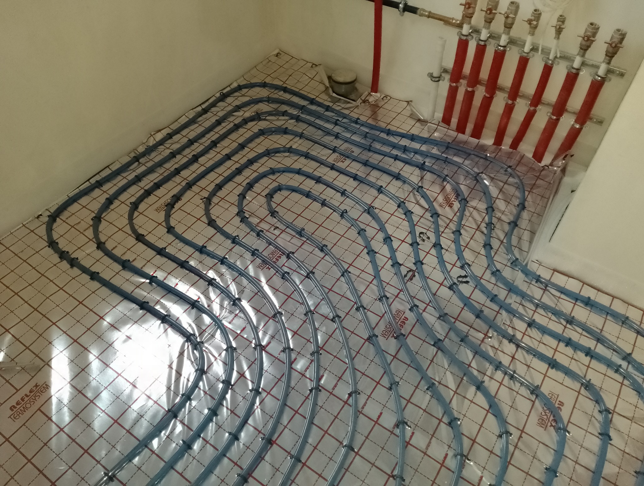 Rozłożone, błękitne rury instalacji grzewczej na folii z nadrukowaną siatką, z widocznymi czerwonymi pionowymi rurami rozdzielacza w tle.