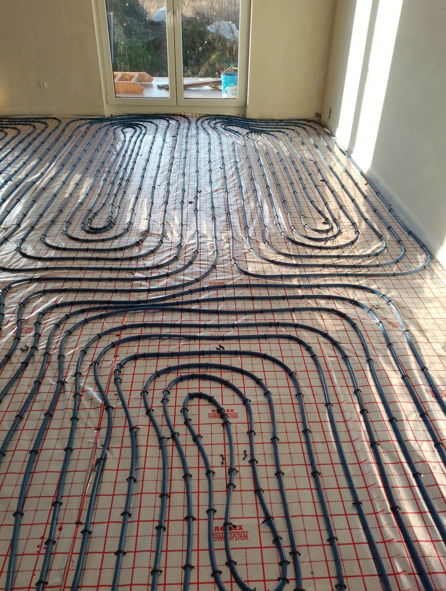 Rury instalacji grzewczej ułożone na podkładzie z siatką w nowym budownictwie, widoczne okno w tle.