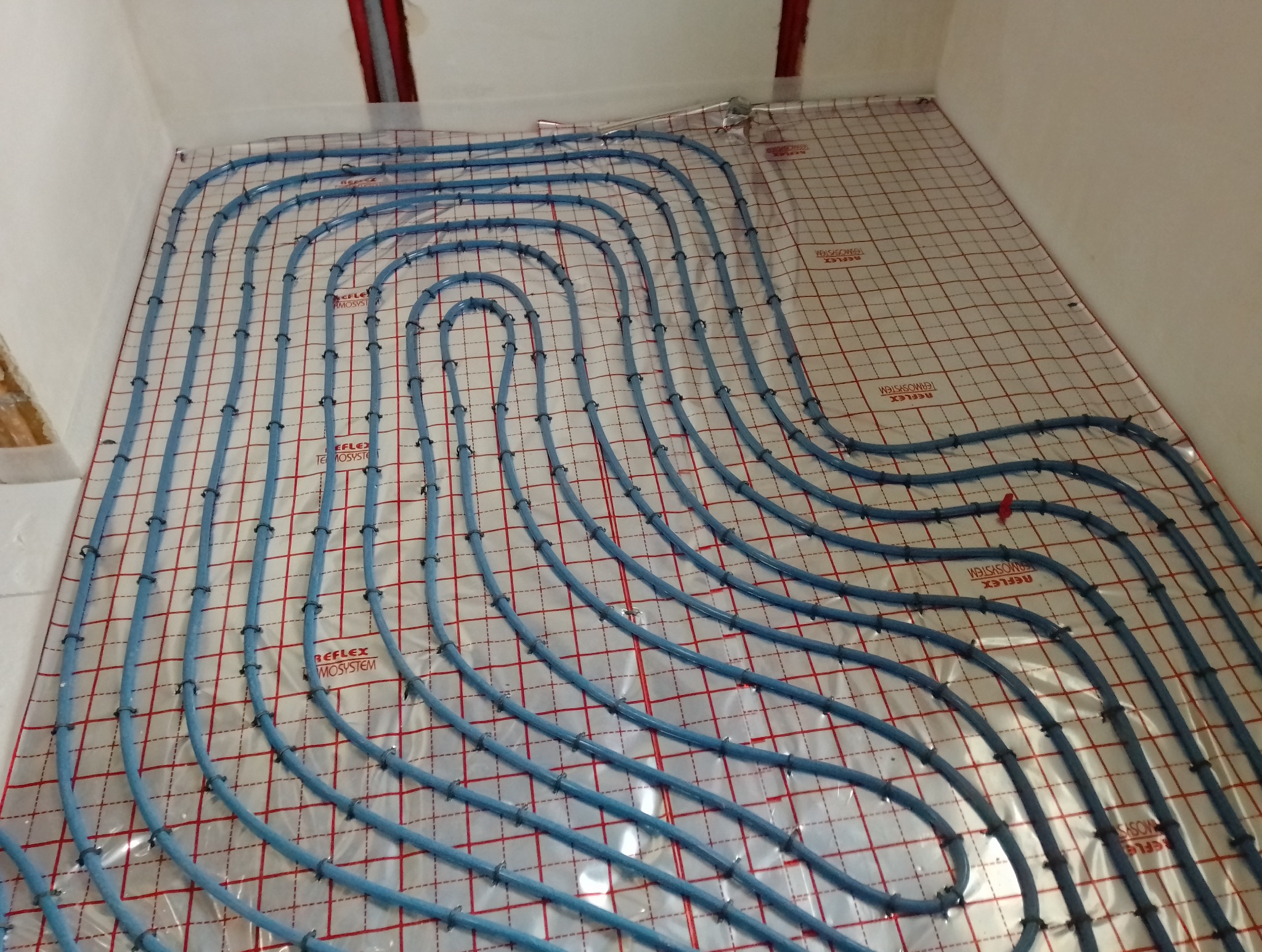 Niebieskie rurki instalacji grzewczej ułożone w pętle na podłodze, przymocowane do folii z czerwoną siatką, widoczne czarne klipsy mocujące i fragment ściany z rurami pionowymi.