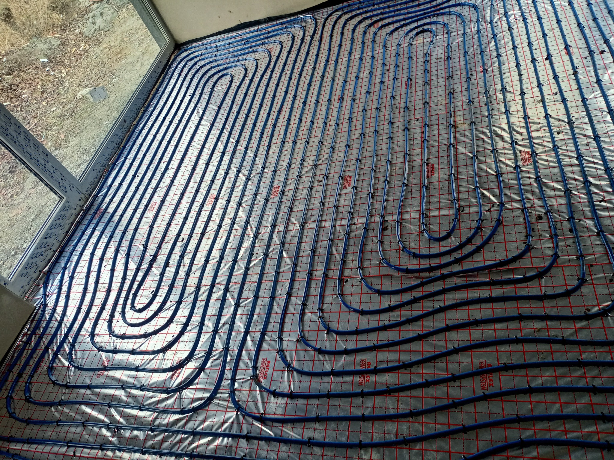 Niebieskie rurki instalacji grzewczej ułożone w pętle na folii z czerwoną siatką, widok z góry, okno w tle.